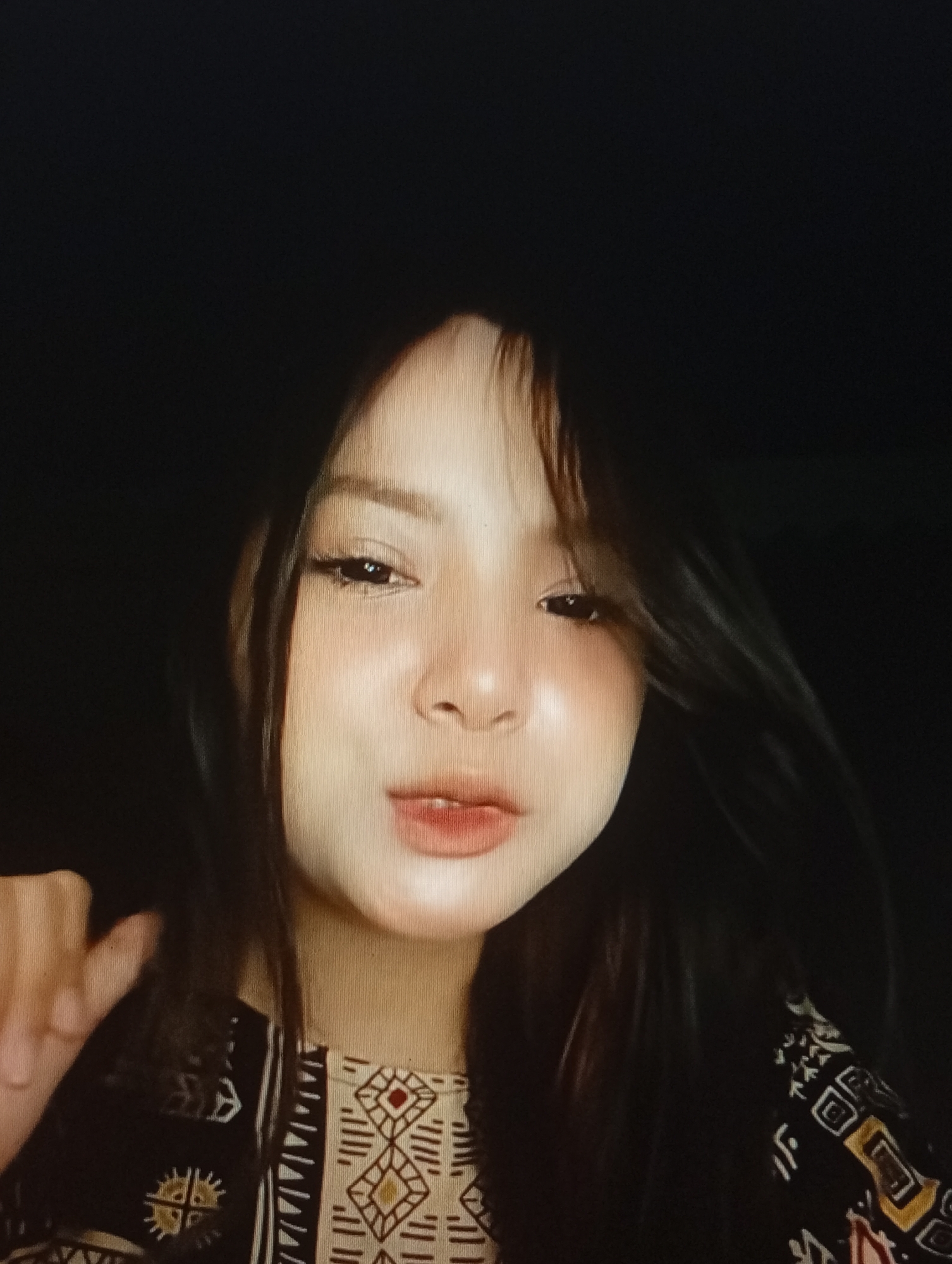 A close-up, low-angle, medium shot features a young woman with dark hair and fair skin against a black background. Her face is illuminated, highlighting her features, including dark eyeliner, blush on her cheeks, and lipstick on her lips. She is wearing a patterned top, visible from the chest down, with a geometric design. Her left hand is visible in the lower left corner, partially obscuring her clothing. The overall lighting is dim, suggesting an indoor setting at night. There is no discernible connection to Lhokseumawe, Indonesia.