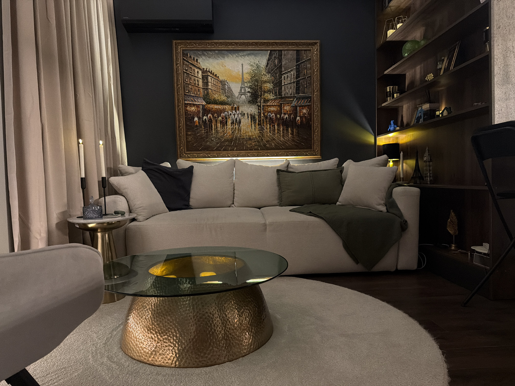 The image depicts a dimly lit, contemporary living room interior, characterized by a dark grey wall and rich brown wooden flooring. Dominating the scene is a large, plush sectional sofa in a neutral beige tone, adorned with several throw pillows in varying shades of beige, charcoal grey, and olive green. A dark olive green throw blanket is casually draped over one section of the sofa.
Above the sofa hangs a framed oil painting of a Parisian street scene, featuring the Eiffel Tower in the background, with pedestrians and shopfronts depicted in a somewhat impressionistic style. To the left of the sofa, a floor-to-ceiling cream-colored curtain is visible, partially obscuring a dark, rectangular object mounted high on the wall, likely an air conditioning unit. Beside the curtain, a small, round side table with a marble top and a gold pedestal base holds two lit white taper candles in black holders and a small blue decorative jar.
In the foreground, a circular, off-white textured rug covers a portion of the wooden floor. Centered on the rug is a modern coffee table with a dark tinted glass top, supported by a striking gold, hammered-metal base.
To the right of the sofa, a dark wooden bookshelf extends from floor to ceiling, filled with various decorative items, books, and some smaller framed pictures. Notable items on the shelves include a blue figurine, several candle holders, and miniature architectural models. A soft yellow light emanates from a lamp on the lowest shelf, casting a warm glow onto the wall and some of the objects. A black metal chair is partially visible in the far right of the frame.
This scene is a living room interior. It is not possible to determine the location within the city of Plovdiv, Bulgaria from the provided image.
