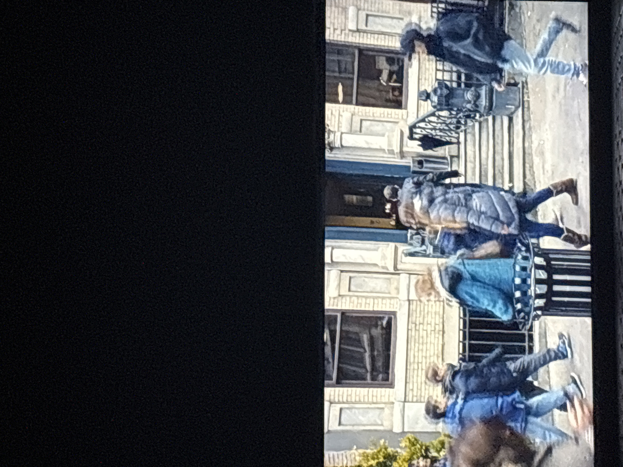 The image displays a bustling urban street scene, likely in Budva, Montenegro, featuring several pedestrians and architectural structures under clear daylight. The photograph itself appears to be a capture of a screen, with a large, unlit black area occupying the left side, and the depicted scene is rotated 90 degrees clockwise.
Focusing on the content of the scene (as if viewed upright):
The street is populated by multiple individuals walking on a paved sidewalk or road. From right to left:
- A person in a dark jacket, light blue jeans, and a dark cap is seen in mid-stride, moving away from the viewer.
- Central to the image, another individual is captured from behind, dressed in a long, light-colored (possibly grey or light blue) quilted puffy coat, dark trousers, and brown boots.
- To their left, part of another person is visible, wearing a light blue jacket or top.
- In the foreground, a cluster of people appears somewhat blurred, with several wearing blue jackets and jeans, suggesting a group moving together.
Lining the street on the left are multi-story buildings, characterized by light-colored facades, numerous windows, and decorative architectural elements such as cornices, which lend a classic European urban aesthetic. Details like a decorative black iron fence or railing and a dark utility box are also present along the street.
The scene is set during the daytime, with bright lighting indicating clear weather. The clothing worn by the pedestrians, including jackets and coats, suggests cool or mild temperatures. The activity primarily involves people walking or gathering on the street. No specific events or discernible emotions are apparent, and there is no visible text within the scene.