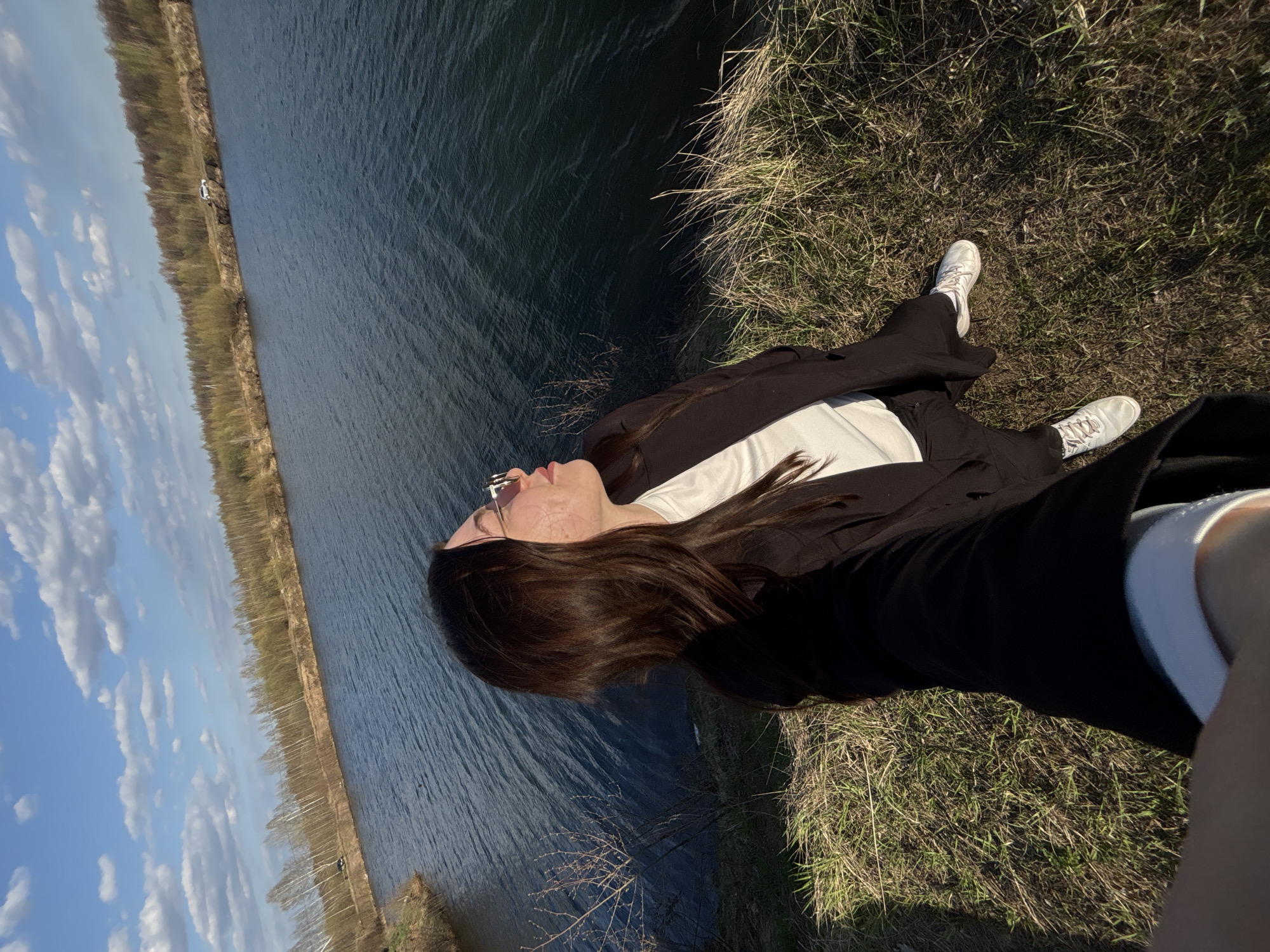 A woman lies on her back on a grassy bank next to a body of water under a blue, partly cloudy sky. The water extends to the left edge of the frame, with a line of trees visible on the far side. The woman is wearing white sneakers, black pants, a white shirt, and a black jacket. She has long brown hair and is wearing sunglasses. The photograph appears to be taken in Zaosinovo, Russia, based on visual details and the presence of a body of water adjacent to a forested area, which is typical of the region.