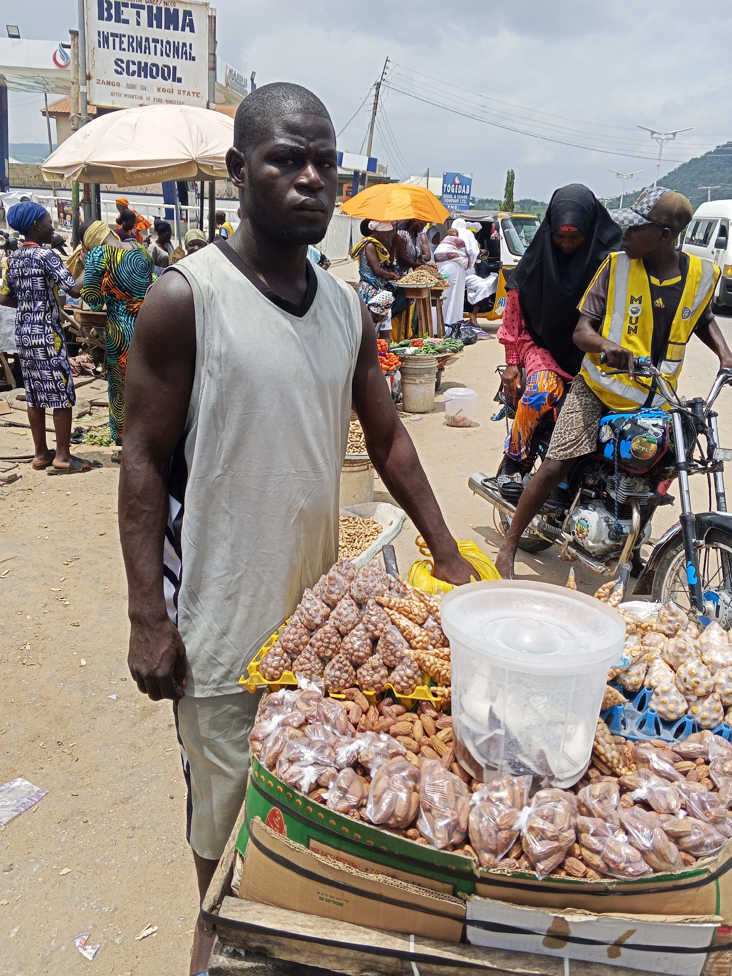 The image depicts a busy street market scene in Zango-ndaji, Nigeria. A man in a light gray sleeveless shirt and light-colored shorts stands in the foreground, looking towards the camera with a neutral expression. He is positioned next to a stall displaying an assortment of nuts and dried fruits, some of which are packaged in clear plastic bags. The stall is made of cardboard boxes and a green-trimmed crate. Behind the man, other people are visible, some dressed in colorful clothing and headscarves. A woman wearing a black hijab and a person in a yellow vest on a motorcycle are in the mid-ground. In the background, a sign for "BETHMA INTERNATIONAL SCHOOL" is prominently displayed. The environment appears to be an outdoor market with a dusty ground, and overhead power lines are visible against a cloudy sky. A few vehicles, including what appears to be a yellow tricycle and a white van, are also present. The overall impression is of a lively and typical market day.
