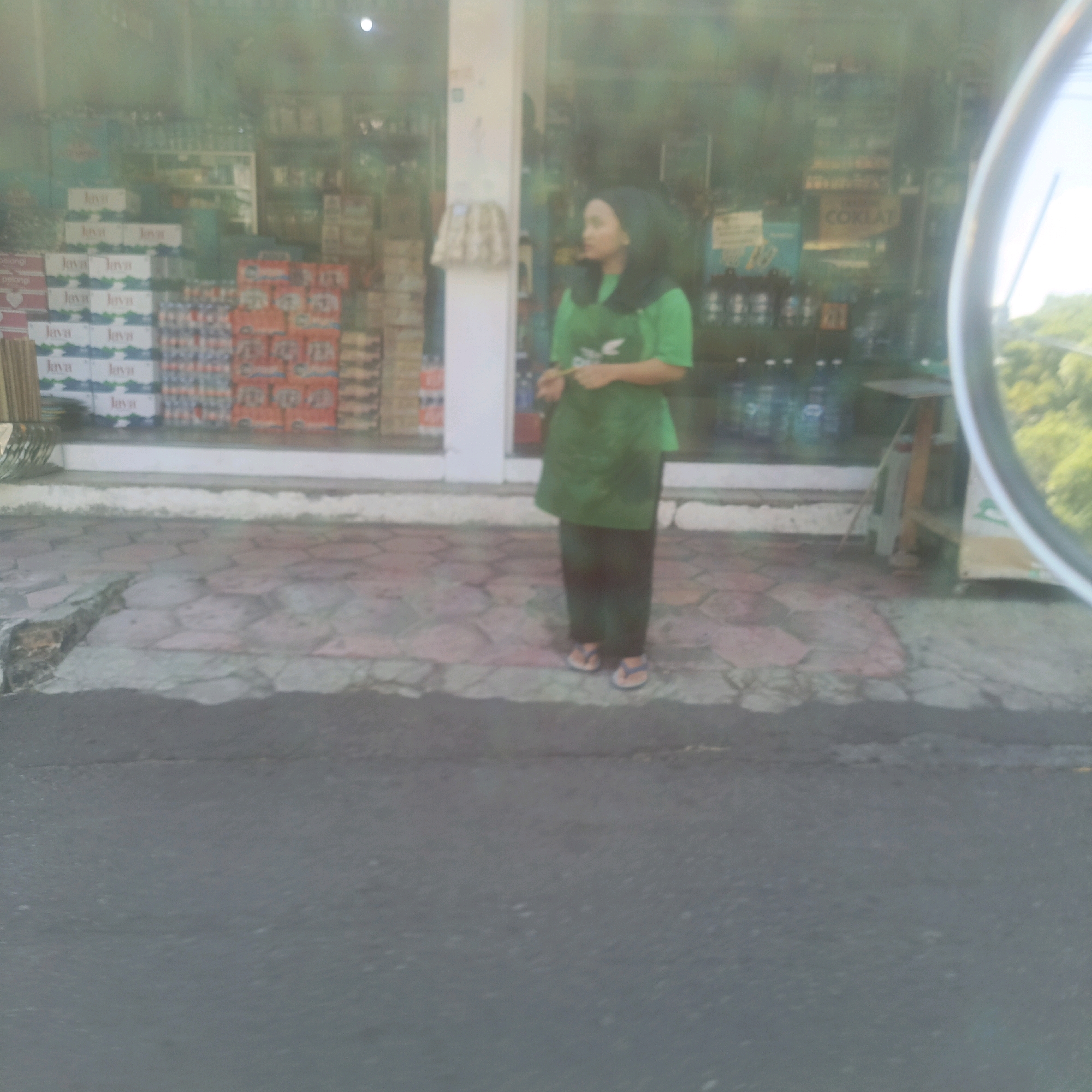 A person stands in front of a store in Salatiga, Indonesia. The person is wearing a green hijab and apron over dark pants and flip-flops. The store has shelves stocked with various beverages, including what appear to be bottled water and soft drinks. The store entrance has a white frame and the floor outside is paved with interlocking hexagonal stones. The image is taken from across a street.
