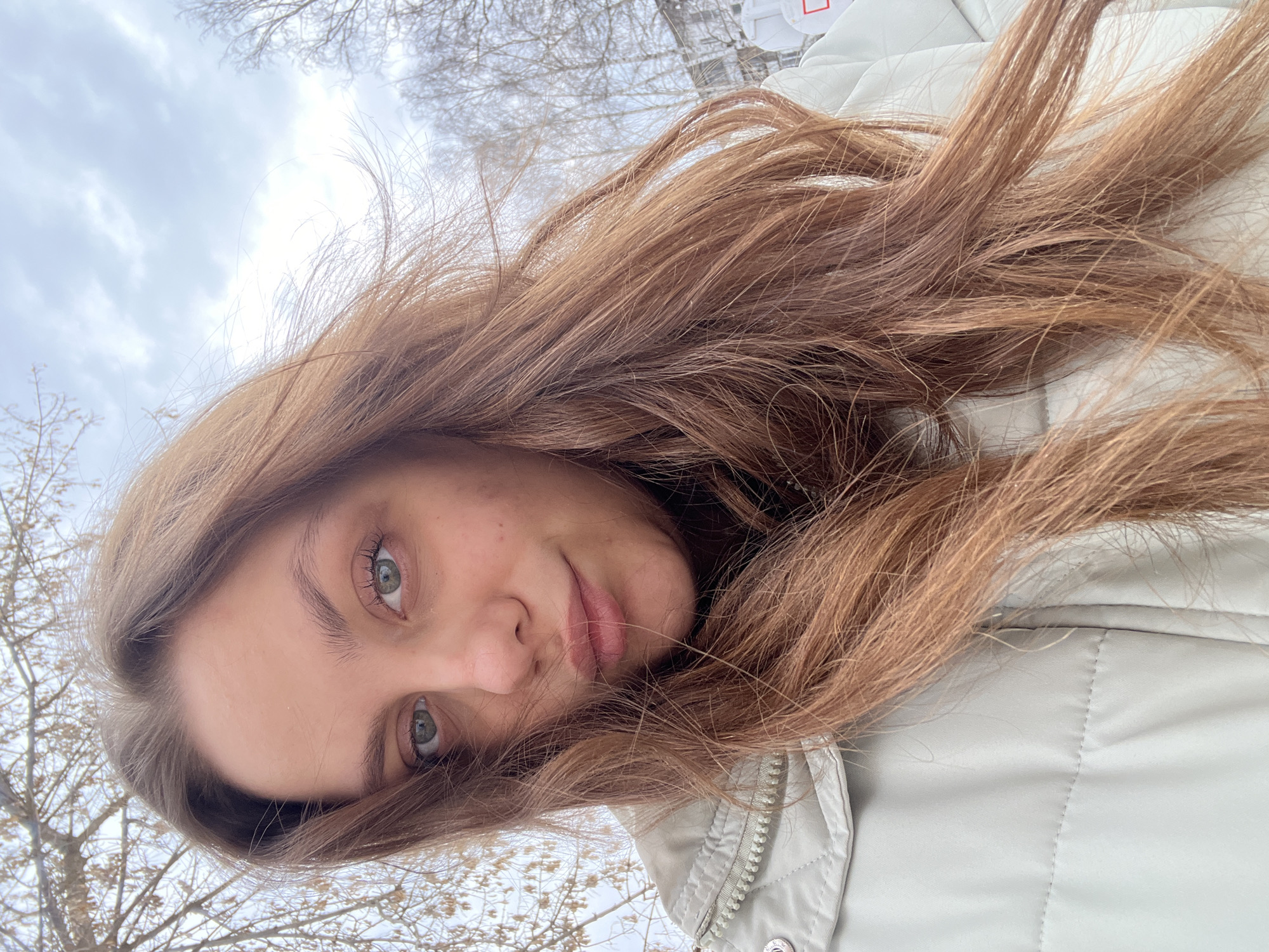 A woman with long, reddish-brown hair is taking a selfie outdoors. She has fair skin and green eyes. She is wearing a light-colored quilted jacket. The background suggests a winter scene with bare trees and a cloudy sky. While the exact location in Chaikovsky, Russia is not discernible from the image, the presence of trees and overcast skies suggests a possibly snowy or cold urban environment.
