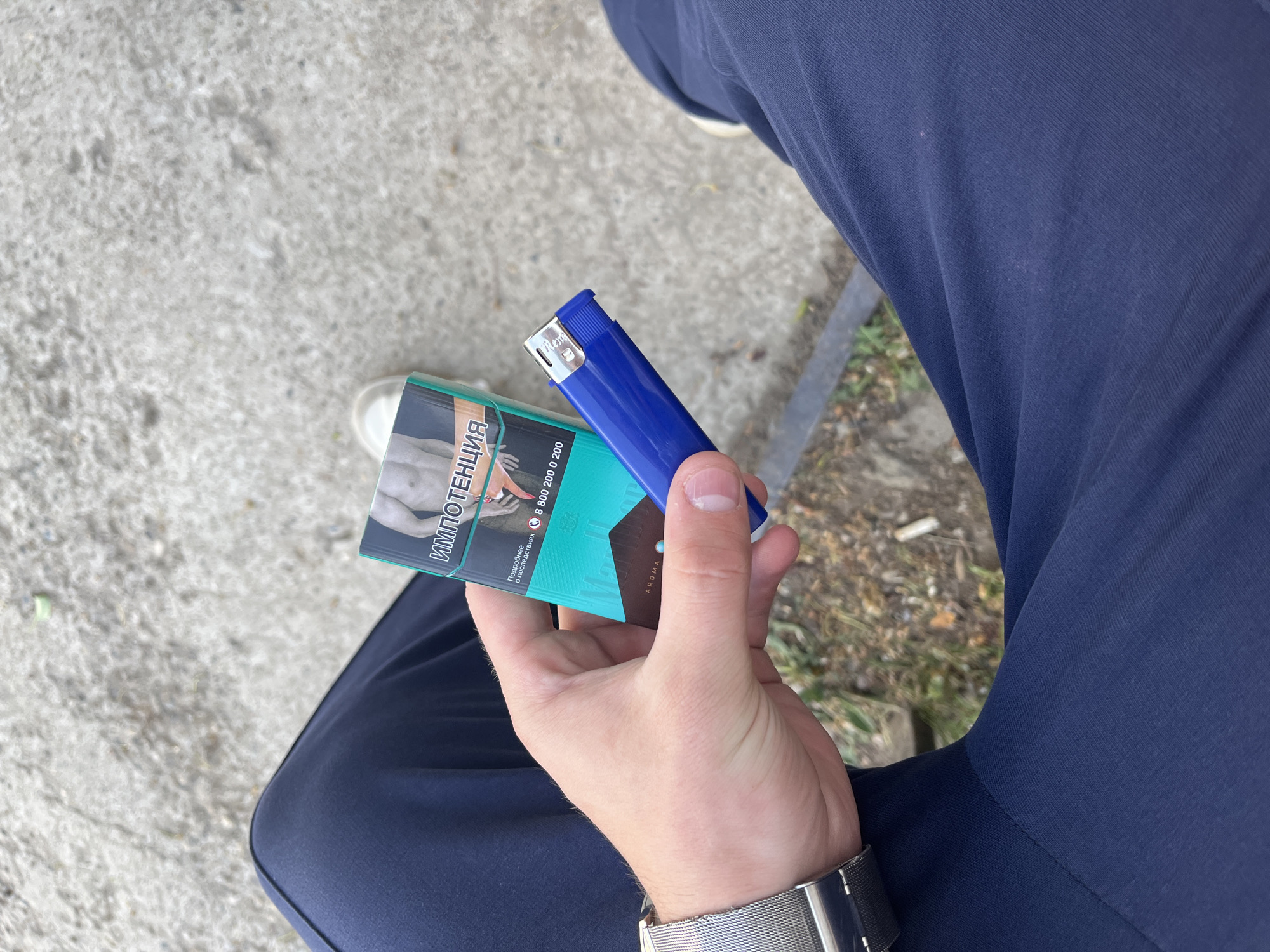 The image shows a person holding a pack of cigarettes and a blue lighter. The cigarette pack has a graphic warning and text in Russian. The person is wearing dark blue trousers and a silver watch. They are sitting outdoors, likely on a bench, with pavement and some vegetation visible in the background. The location is likely Chaikovsky, Russia, based on the language on the cigarette pack and the presence of elements typical of a Russian urban environment.