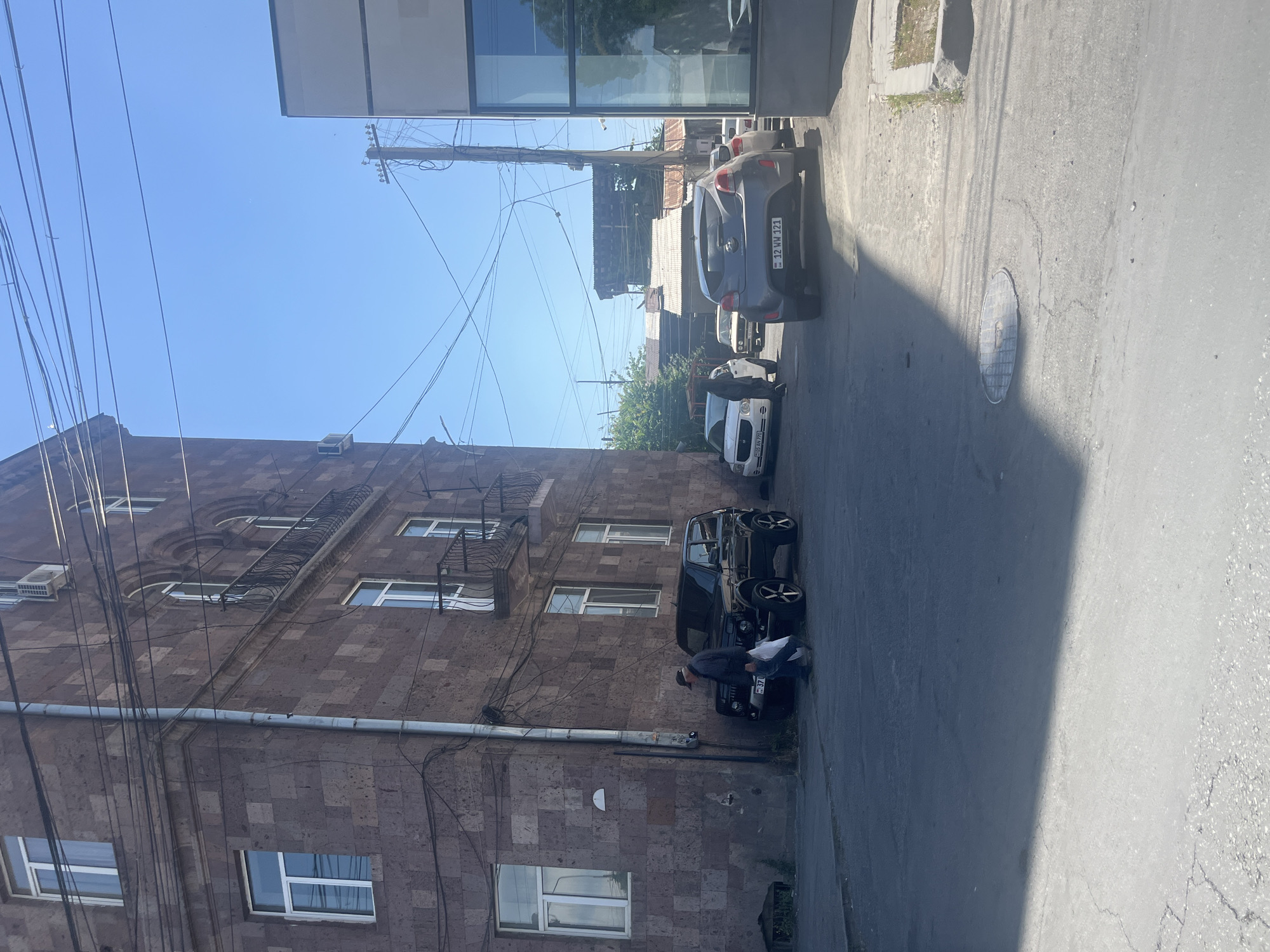 The image captures a sunlit street scene in what appears to be an urban setting, likely Yerevan, Armenia, given the architectural style and visible license plates. The sky is bright blue and clear, indicating a sunny day, possibly morning or early afternoon, as evidenced by the distinct shadows cast on the asphalt road.
On the left, a multi-story building constructed from reddish-brown stone, characteristic of Armenian architecture, dominates the frame. It features numerous windows, some with small wrought-iron balconies. A dense network of black utility cables and a grey pipe run across its facade. On the right, a modern building with a large reflective glass facade is partially visible.
Several cars are parked along the street. In the foreground, a black SUV or Jeep-like vehicle with distinctive black custom rims is parked next to the stone building. A person wearing a dark cap and jacket is leaning over the front of this black car, seemingly inspecting or working on it. Further down the street, a white compact car is partially visible, followed by a grey hatchback.
Visible text includes license plates on two of the cars. The grey hatchback displays the plate "12 HW 121". A partially visible plate on the white car appears to be "05 AN 020", and a plate on the black car partially shows "37 ZW". Overhead, a tangle of electrical wires crisscrosses the sky, emanating from a utility pole. A circular metal manhole cover is visible on the street surface. The overall impression is that of a bustling, lived-in city street.