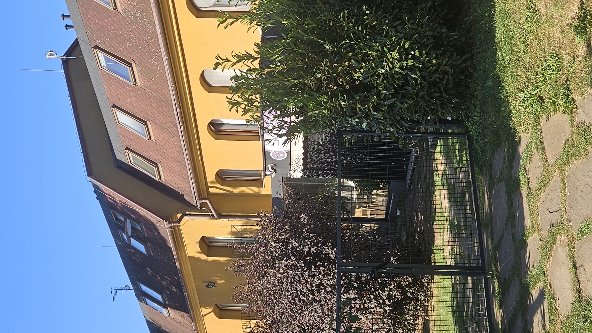 The image shows a yellow building with a red tiled roof and several dormer windows, located in Prešov, Slovakia. In front of the building is a black metal fence and a path made of irregularly shaped stone slabs interspersed with grass. To the left of the path, a dark purple, bushy tree stands, and to the right, a dense green hedge. Through the fence, part of a courtyard with tables and chairs can be seen. A sign with a circular logo featuring a stylized ice cream cone is visible on the building, suggesting a business establishment. The sky is clear blue, and the sunlight casts shadows. No people are visible in the image.