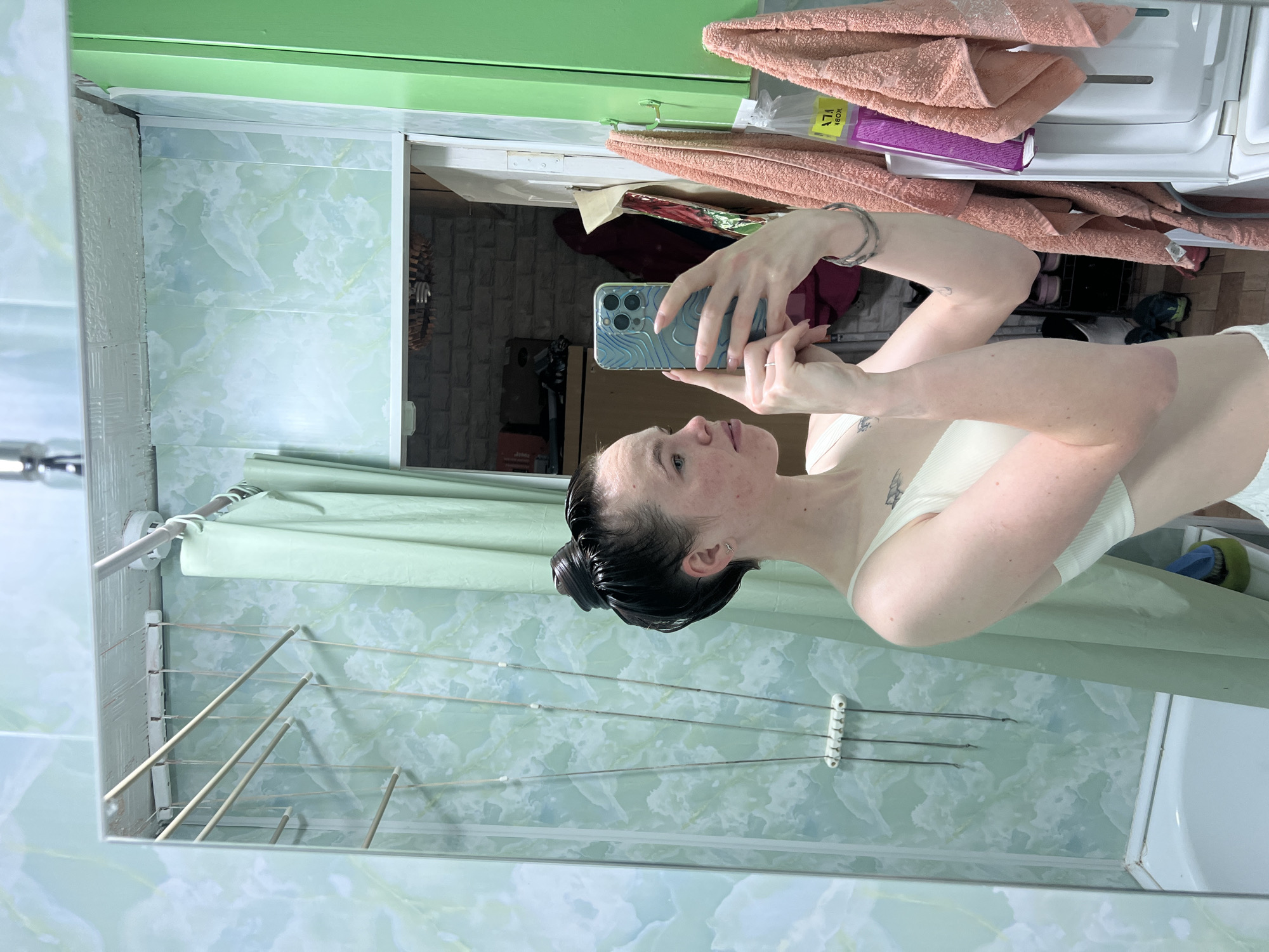 A woman takes a selfie in a bathroom. The bathroom has light green-and-white patterned tile walls and a green trim. The woman is holding a phone with a light blue patterned case. She is wearing a light-colored top. Her hair is pulled back. A clothes-drying rack is attached to the wall. A white cabinet with peach-colored towels on top is in the background. Through an open doorway, a room with exposed brick can be seen. The location is Chaikovsky, Russia.
