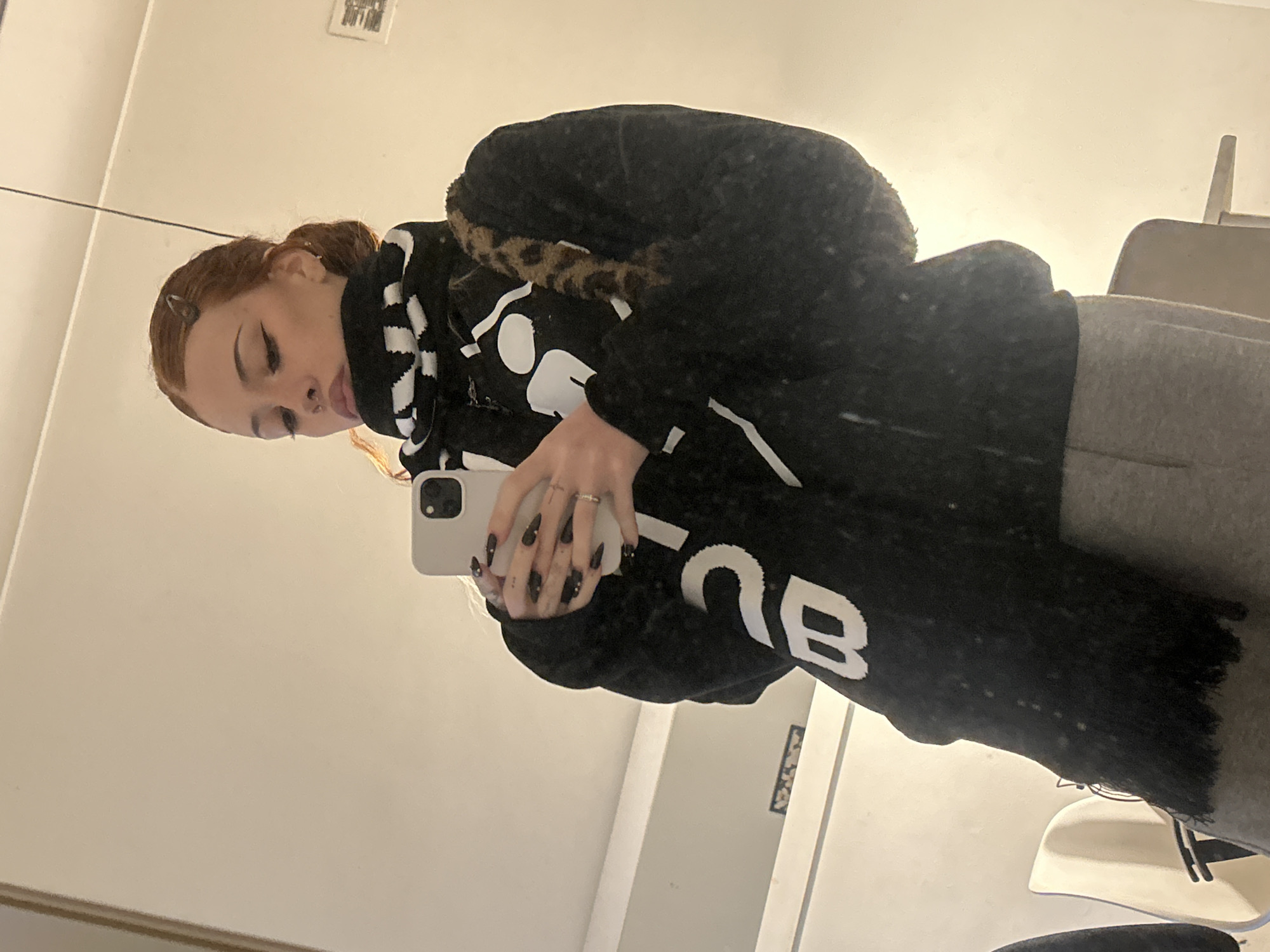 A young woman with auburn hair styled in a bun, wearing a black fuzzy hooded sweatshirt with white lettering and a black and white checkered scarf, is taking a selfie in a mirror. She has bold eye makeup and long, pointed black nails. She is also wearing grey sweatpants. A small leopard print furry bag is slung over her shoulder. The background appears to be an indoor space with white walls and furniture reflected in the mirror. There is no identifiable information in the image that places the scene in Buenos Aires, Argentina.