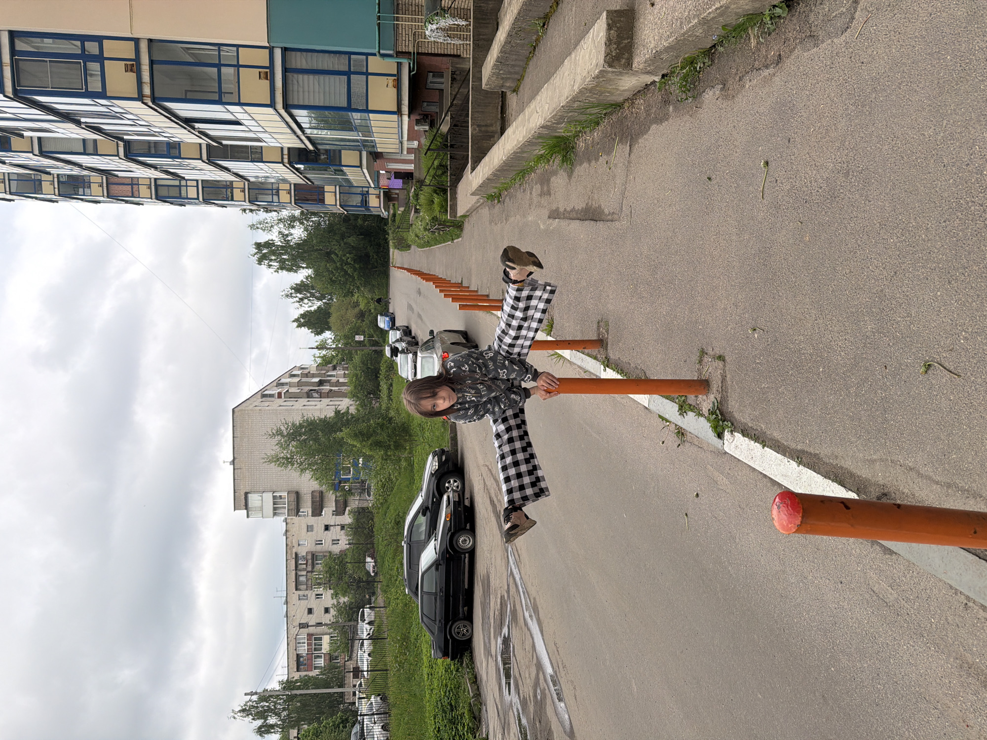 A child sits on an orange barrier along an asphalt road in Tokkari-Lend, Russia. The child wears black and white checkered pants and a grey patterned jacket. A row of orange barriers lines the road. Several cars are parked along the road behind the child. Buildings are visible in the background. The sky is overcast.