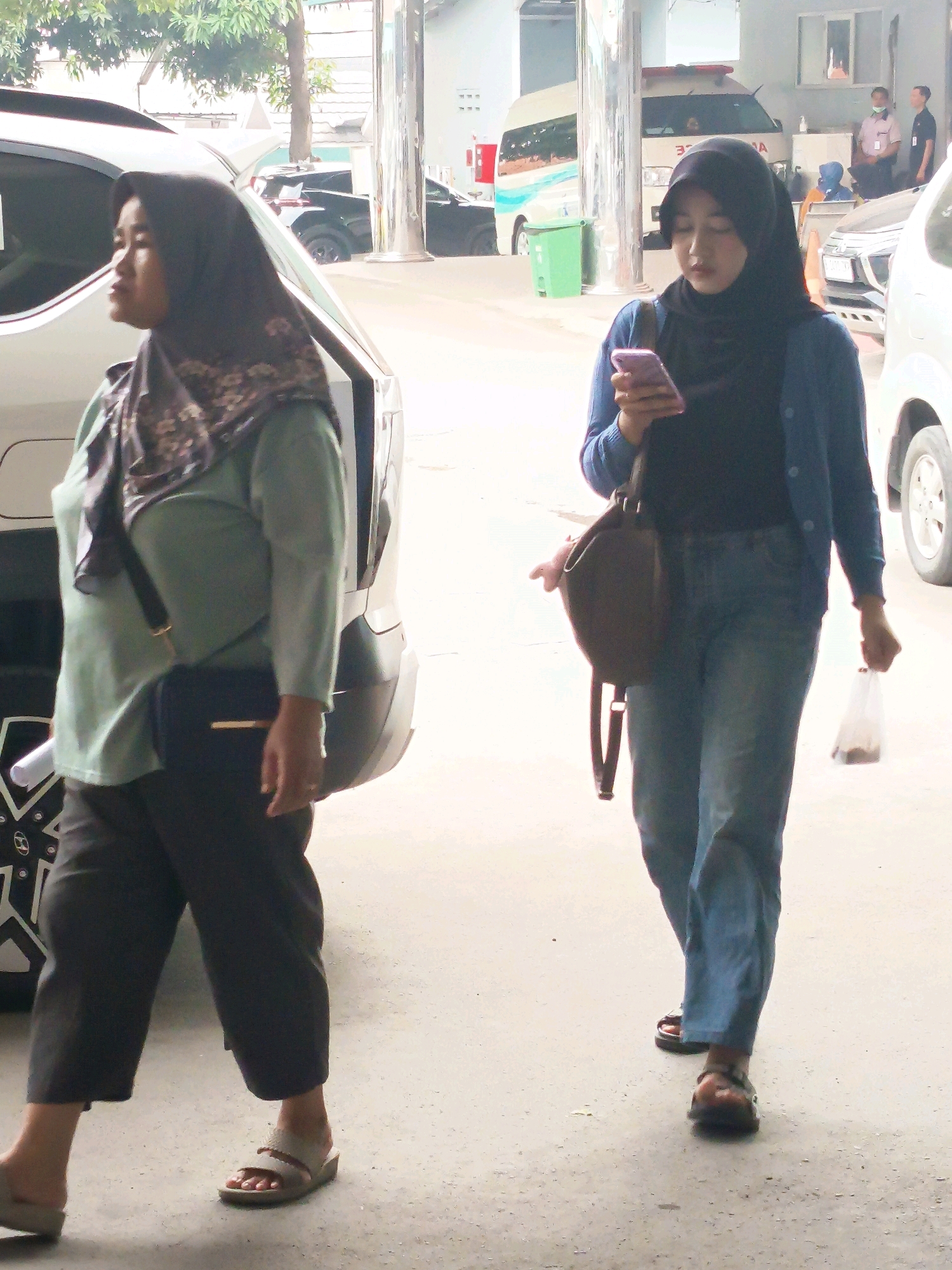 Two women are walking in an outdoor, paved area, likely in Tangerang, Indonesia, given the context. The woman on the left wears a muted green long-sleeved shirt, dark pants, a dark purse slung across her body, and light-colored slip-on sandals. She is also wearing a patterned headscarf. The woman on the right is wearing a dark top, a blue cardigan, blue jeans, and dark sandals. She is holding a smartphone in her right hand and a small plastic bag containing an item in her left hand. She has a brown shoulder bag. In the background, there are vehicles, including what appears to be an ambulance, and a green trash bin. The environment suggests a public or service area, possibly near a building.