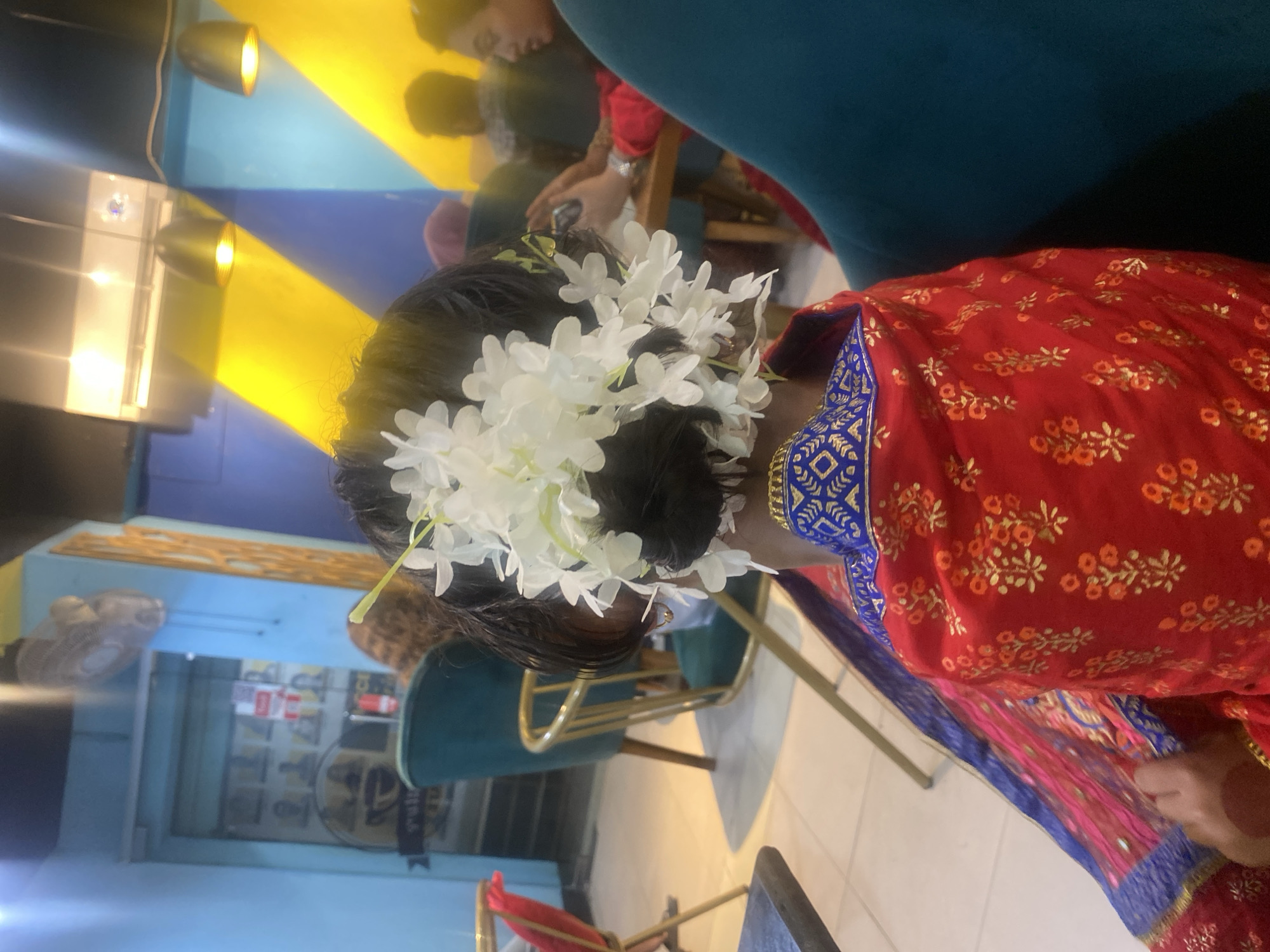 A woman with white flowers in her hair wears a red and blue sari. She sits indoors in a blue chair. Other people are in the background. The scene is in Dhaka, Bangladesh.