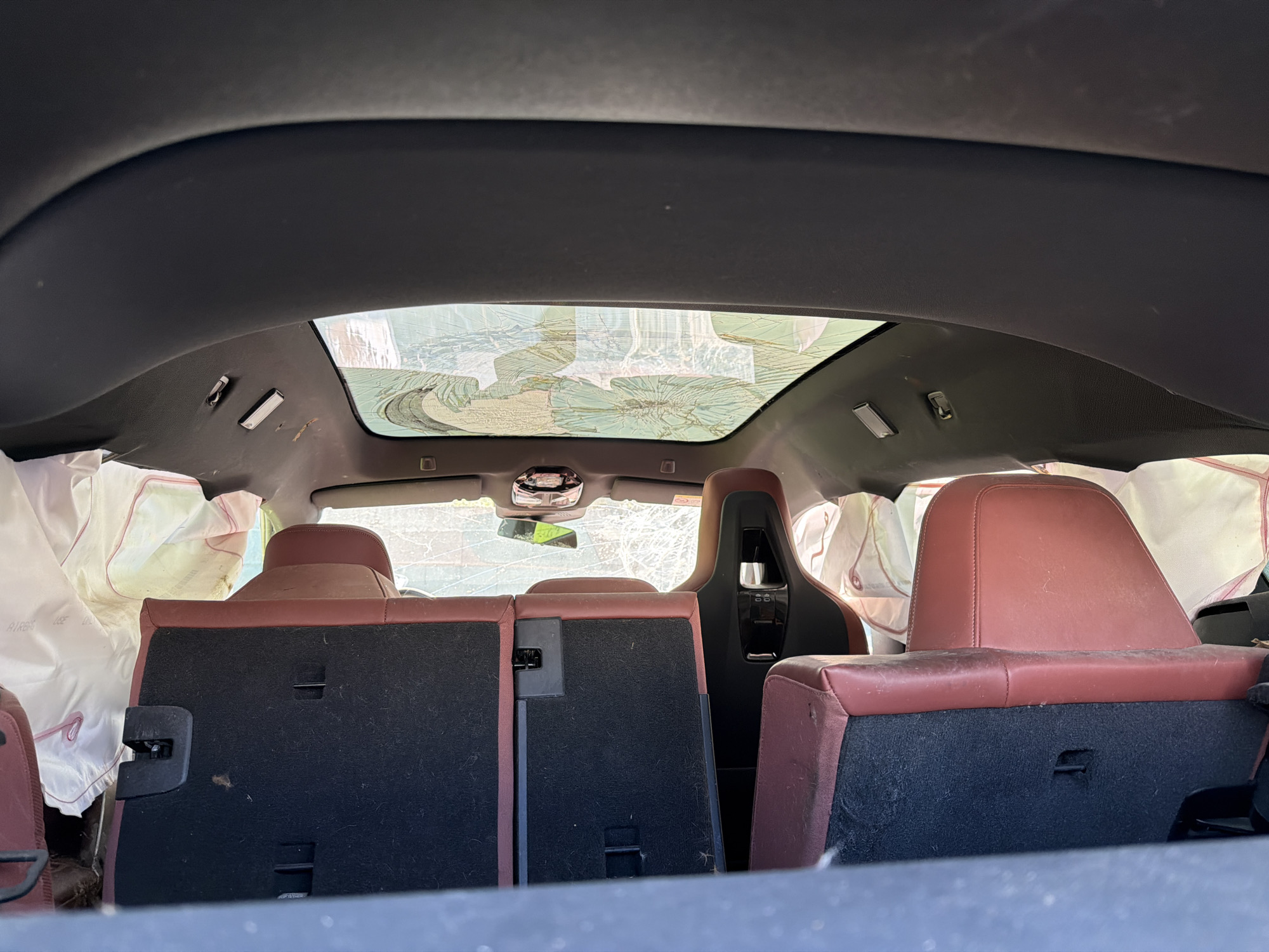 The image shows the interior of a damaged car, likely after an accident. The panoramic sunroof is shattered. Airbags have been deployed in the back seat. The rear seats are a reddish-brown color. The location is not discernible from the image. There are no people visible.