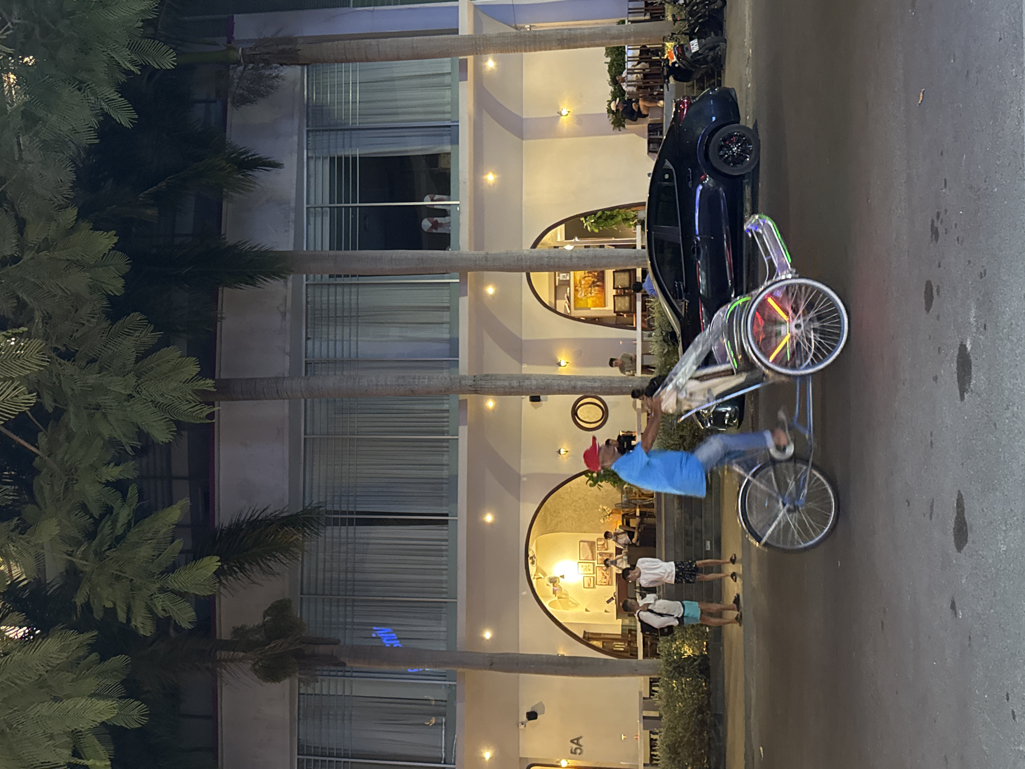In Nha Trang, Vietnam, at night, a man in a blue shirt and red cap is riding a cyclopousse, a three-wheeled vehicle for passengers, down a street. The cyclopousse has illuminated wheels and is in motion. In the background, a modern building with large windows and arched entrances is visible. Palm trees line the street in front of the building. Several people are seen near the building's entrance, some seated and others standing. A dark-colored car is parked on the street next to the cyclopousse, and motorcycles are parked further down the street. The overall scene depicts a typical street view in a Vietnamese city at dusk or nighttime, with a mix of pedestrians, vehicles, and commercial establishments.