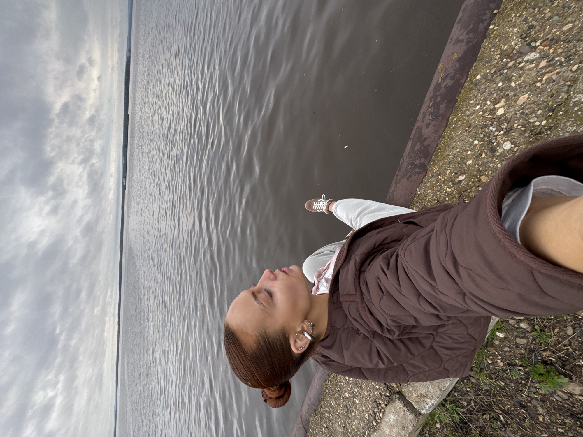 The image depicts a woman sitting on a rocky embankment or pier adjacent to a body of water, likely the Kama River in Chastyye, Russia, given the provided location. The sky is overcast. The woman wears a brown quilted jacket, light-colored trousers, and brown shoes. Her hair is styled in a bun. She appears to be looking out at the water. The water has small ripples and is dark in color. A rusty metal pipe runs along the edge of the embankment. The ground near the woman is covered in small stones and some sparse vegetation.