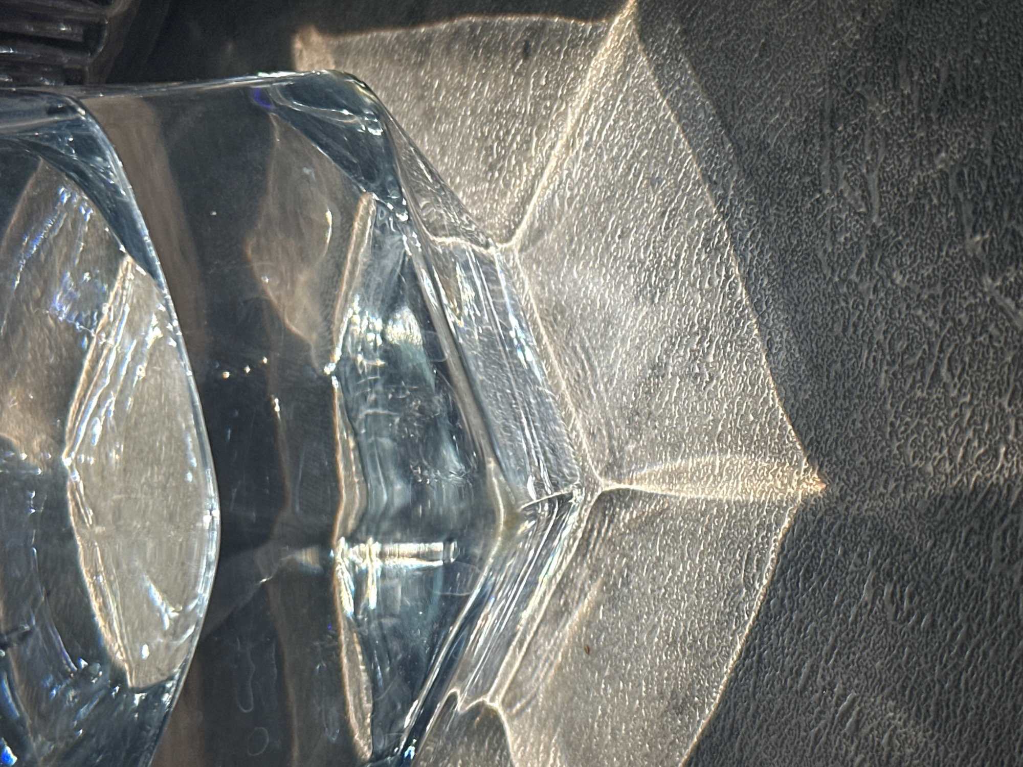 An indoor scene, set in Kazan, Russia, features a transparent, multi-faceted container resting on a dark, textured surface. The container, appearing to be glass or rigid plastic, is partially filled with a clear liquid. Direct light, consistent with sunlight, illuminates the scene from an implied upper-left source. This light interacts with the container and liquid, creating distinct refractive patterns and bright reflections on the textured surface below and around the container's base. These patterns include elongated, angular shapes of intense light. Portions of the textured surface, characterized by fine, irregular bumps, are in shadow. No persons or other specific contextual elements are present.