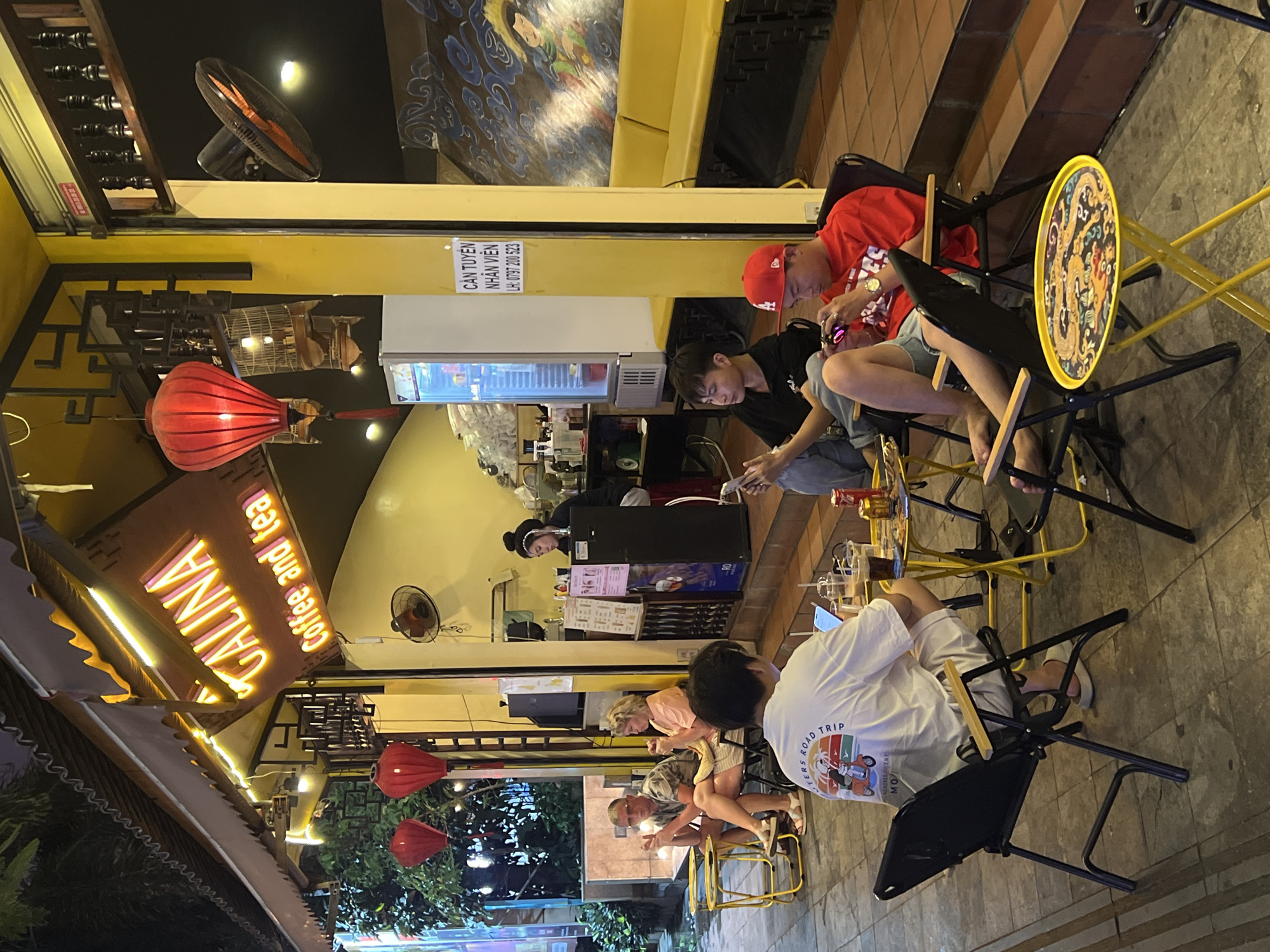 The photograph depicts an outdoor seating area of "GALINA Coffee and tea," located in Nha Trang Ward, Vietnam. Several groups of people are seated at small tables with yellow, folding chairs. Most are looking at their phones or electronic devices. A neon sign displays the cafe's name. Red lanterns hang above. A refrigerator with drinks and a cashier area are in the background. A sign reads "CẦN TUYÊN NHÂN VIÊN LH: 0797 200 523." A fan is attached to the ceiling.
