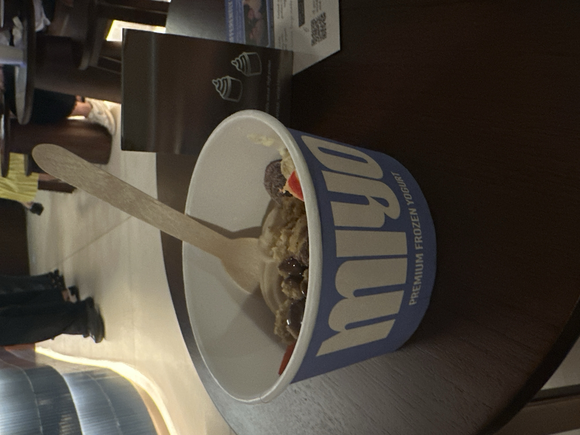 The image displays a cup of Miyo Premium Frozen Yogurt, placed on a dark wooden table in an indoor commercial setting. The white paper cup prominently features a blue logo with the partially visible text "Miyo" and "PREMIUM FROZEN YOGURT". Inside the cup, light-colored frozen yogurt is adorned with various toppings, including what appear to be dark berries, a piece of red fruit, and possibly cookie crumbles. A light-colored wooden spoon is inserted into the yogurt, suggesting it is either being consumed or has just been prepared.
In the background, a dark brown stand or display board is visible, featuring white outline graphics of two cupcakes. Behind this stand, a piece of paper with unreadable text and a QR code is partially obscured. The wider background is blurred, revealing elements of a public indoor area, possibly a food court or shopping mall. Glimpses of other tables and chairs can be seen, along with the lower bodies of two individuals in the far left foreground. One person is wearing dark pants and shoes, standing near a curved architectural element with warm lighting that resembles an escalator. Another person wearing a bright yellow shirt is partially visible further back on the left. The lighting is artificial, typical of an indoor environment. The scene captures a casual moment in a public space, consistent with locations like Mengwi, Indonesia, where such frozen yogurt establishments are common.