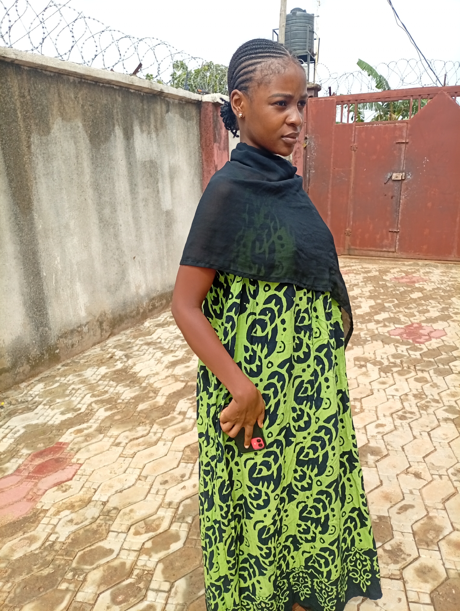 A woman stands outdoors in Unknown, Unknown, facing slightly to the right. She has dark skin and her hair is styled in braids. She is wearing a long, flowing green dress with black patterns, and a black shawl is draped over her shoulders. She holds a phone in her right hand. The background includes a gray concrete wall topped with barbed wire on the left, and a red metal gate on the right. A water tank is visible above the gate. The ground is paved with patterned tiles.