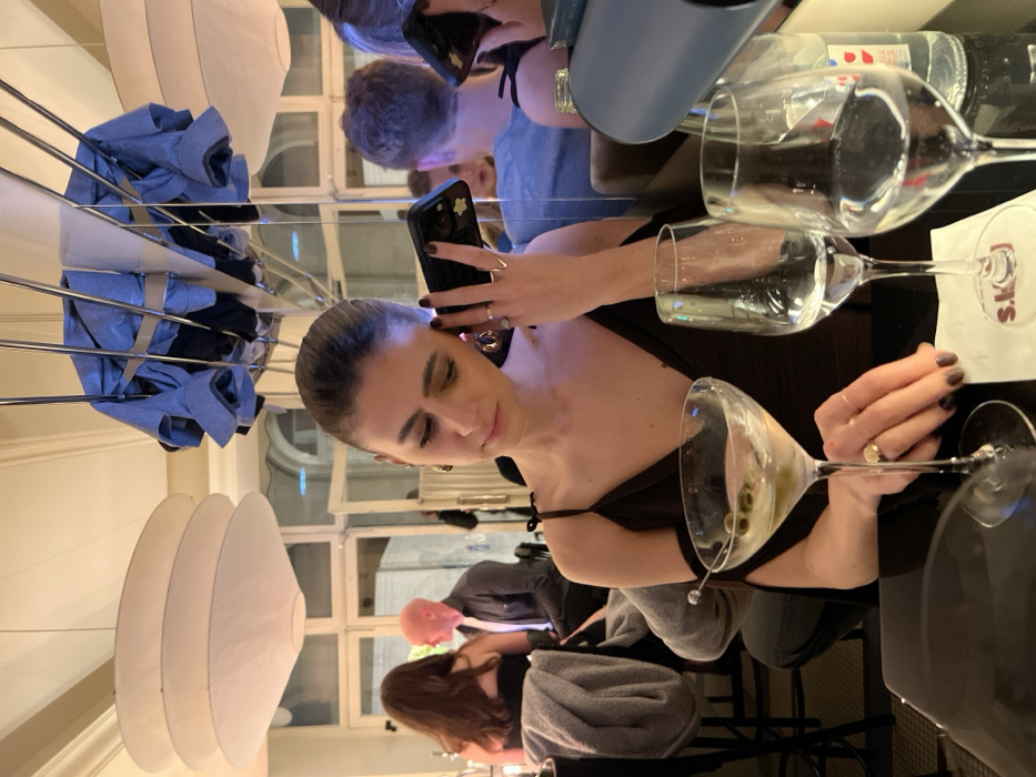 A woman in a black dress takes a photo with her phone. She is seated at a table with drinks, including a martini. Other people are visible in the background. The setting appears to be a restaurant with white hanging lights. The image is shot at an unusual angle, with the woman positioned sideways in the frame.