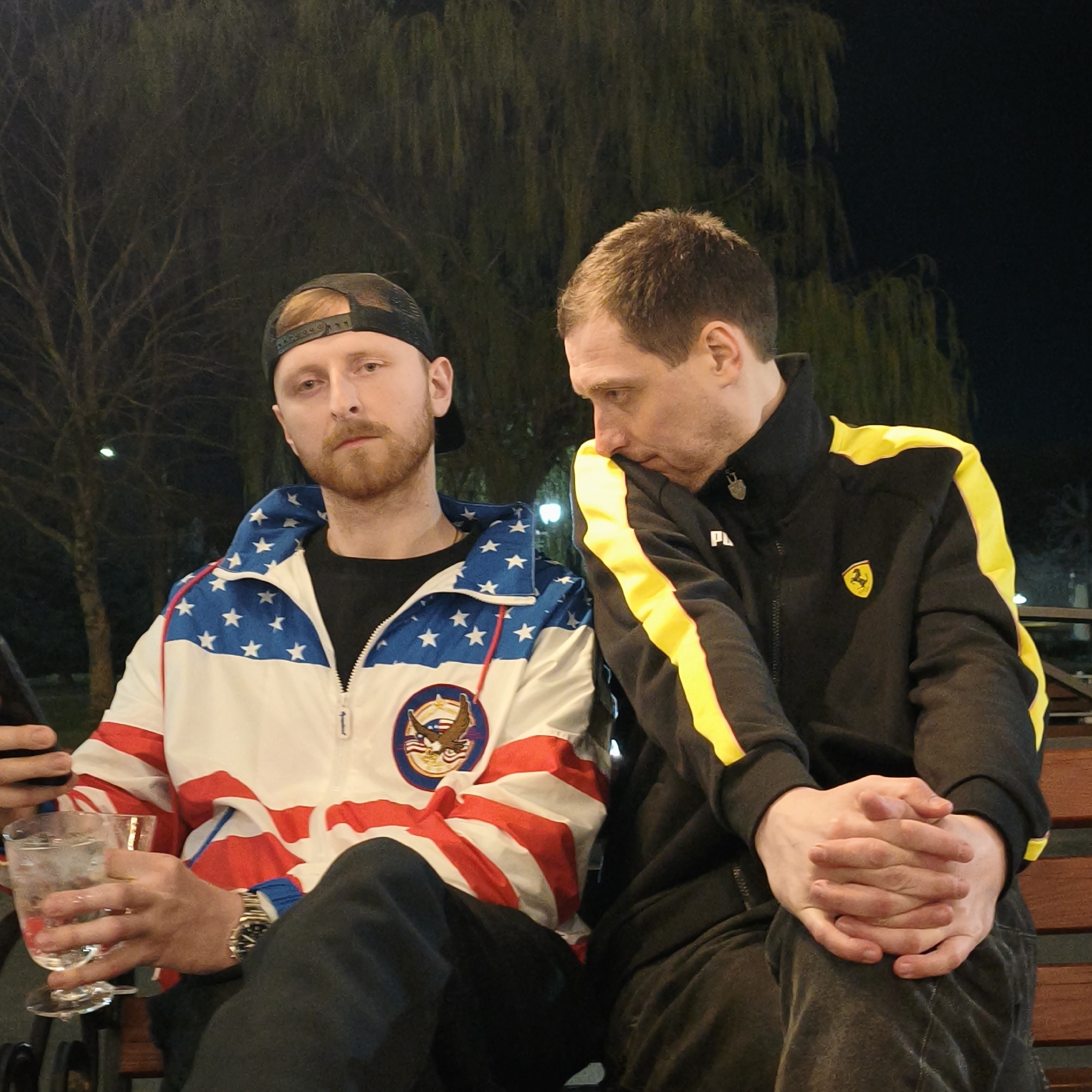 Two men are sitting on a wooden bench outdoors at night. The man on the left wears a black baseball cap, an American flag-themed zip-up jacket, black pants, and a watch. He is holding a clear glass with liquid and a phone. The man on the right wears a black jacket with yellow stripes and a Ferrari logo, and black pants. His hands are clasped. Trees are visible in the background. It is not possible to identify the location in Hrodna.