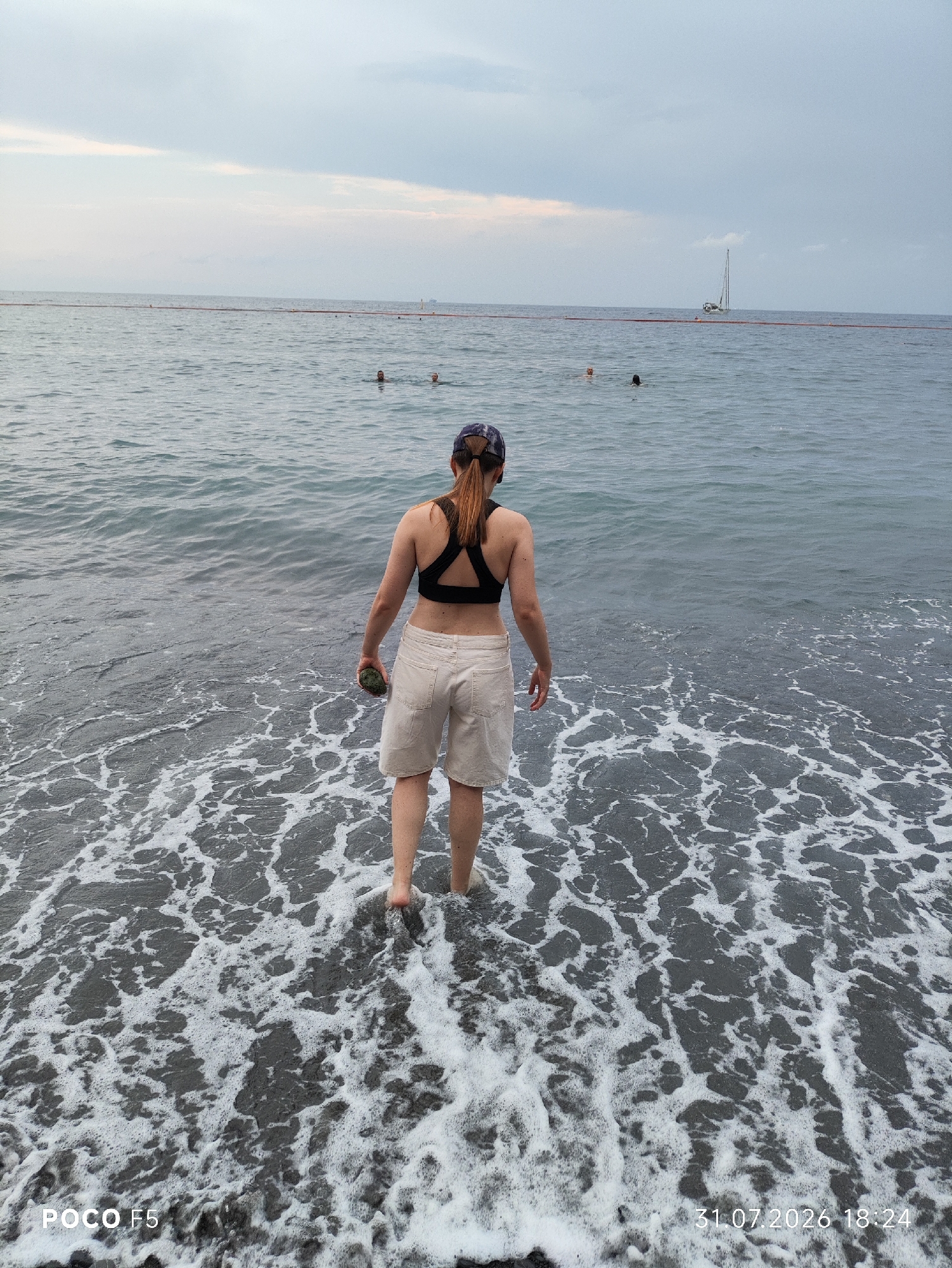 A person stands at the edge of the sea, their back to the camera, with waves lapping at their feet. They are wearing a black sports bra and light-colored shorts, and a baseball cap is on their head. They are holding a small, dark object in their right hand. The sea is choppy, with white foam from the waves creating a textured pattern on the dark sand. In the distance, several people are swimming in the water, and a sailboat is visible on the horizon. The sky is overcast with hints of pink near the horizon. The timestamp "31.07.2026 18:24" and "POCO F5" are present in the lower corners of the image.