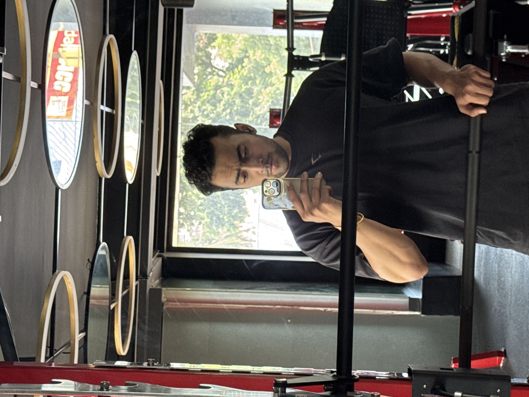 A man with curly dark hair and a beard is taking a mirror selfie in a gym. He is wearing a black t-shirt and has a gold bracelet on his left wrist. He is holding a smartphone with a gold case in his right hand. Behind him, there is a window with trees visible outside, and in the ceiling, there are modern circular light fixtures. The gym equipment, including a barbell and a squat rack, is visible in the foreground and background. The environment suggests an indoor fitness facility. This scene does not provide any specific identifiable landmarks or context to pinpoint its location within Kolkata, India.
