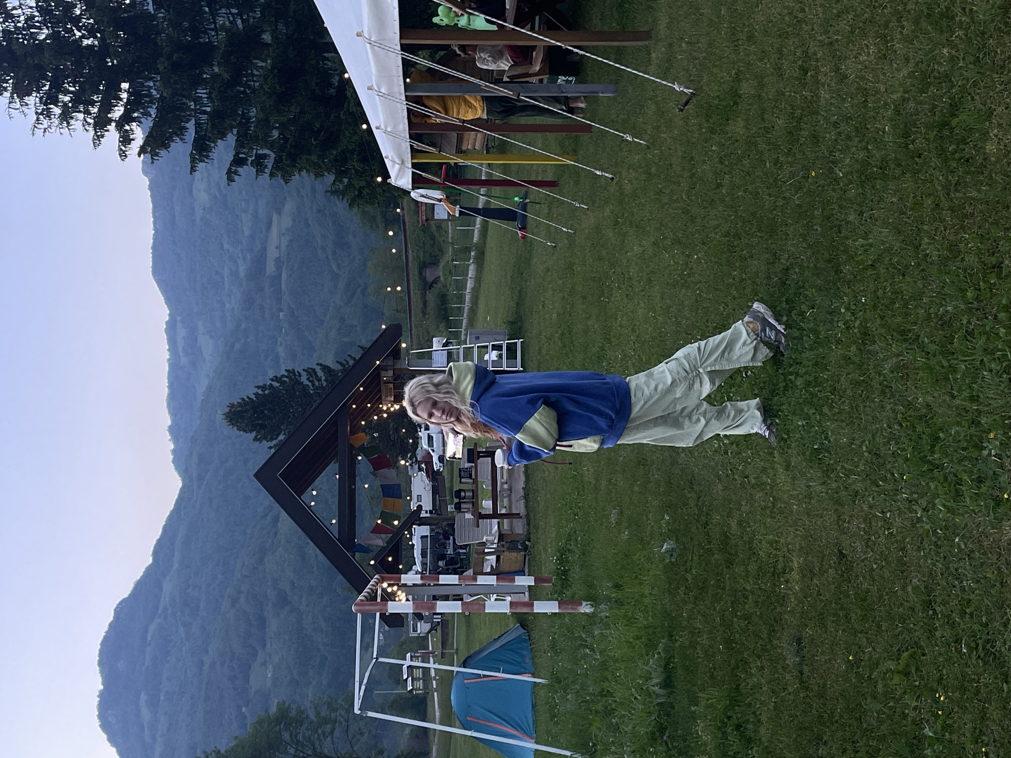 The image depicts a young woman standing on a grassy field in Polja, Montenegro. She is facing the camera, wearing a blue and lime green hoodie and light green pants. Behind her, there is a mountain range, a small building decorated with lights and colorful flags, and camping equipment. A tent is visible in the lower left corner. The scene appears to be at dusk or early evening.