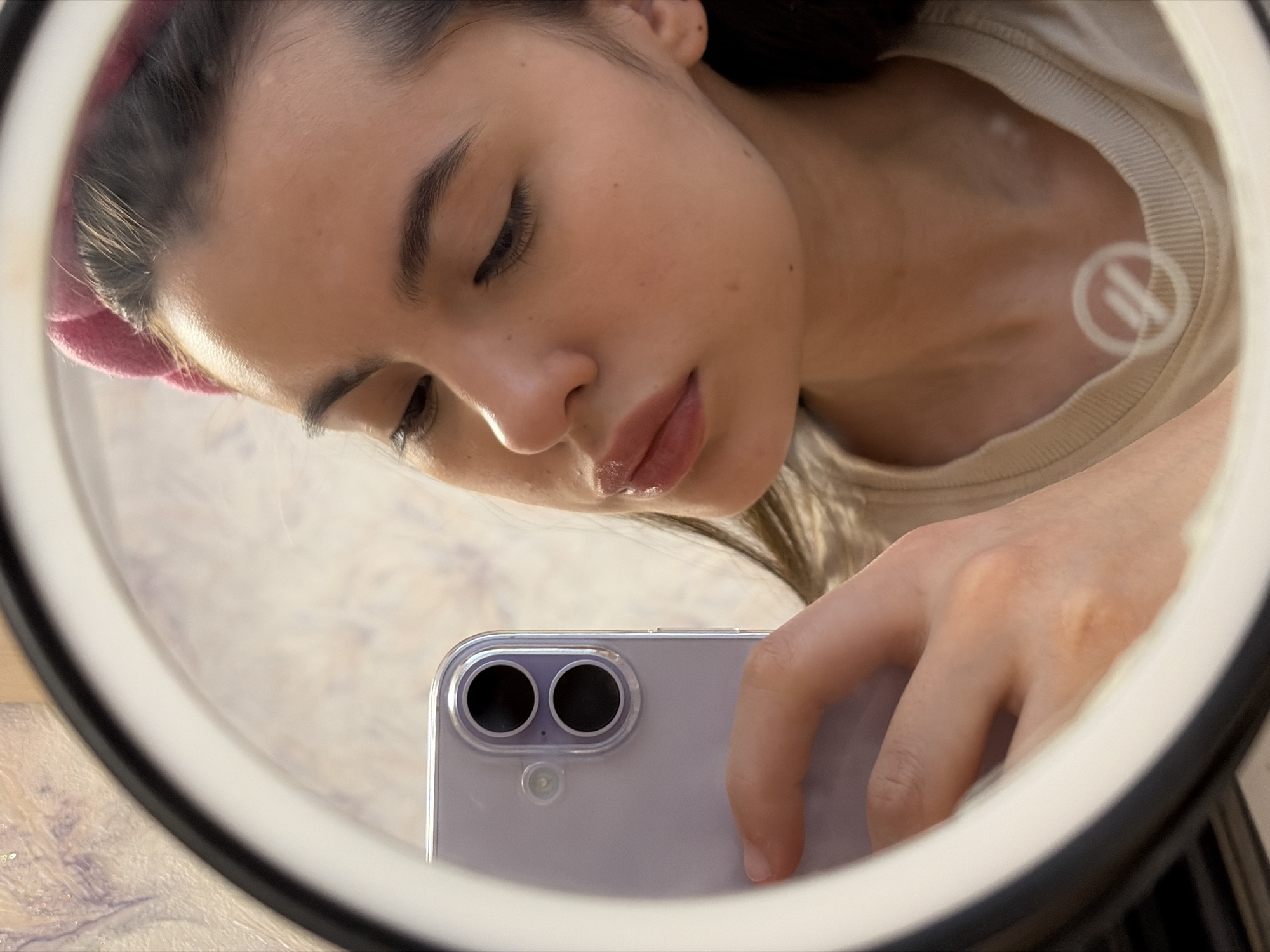 A young woman is holding a smartphone in front of her face, reflected in a ring light. The smartphone has a dual-lens camera system on its back. The woman has fair skin, dark hair partially covered by a pink headband, and glossy lips. She is wearing a light-colored t-shirt with a white logo on the collar. The background is slightly blurred. There is no indication this photo was taken in Chaikovsky, Russia.