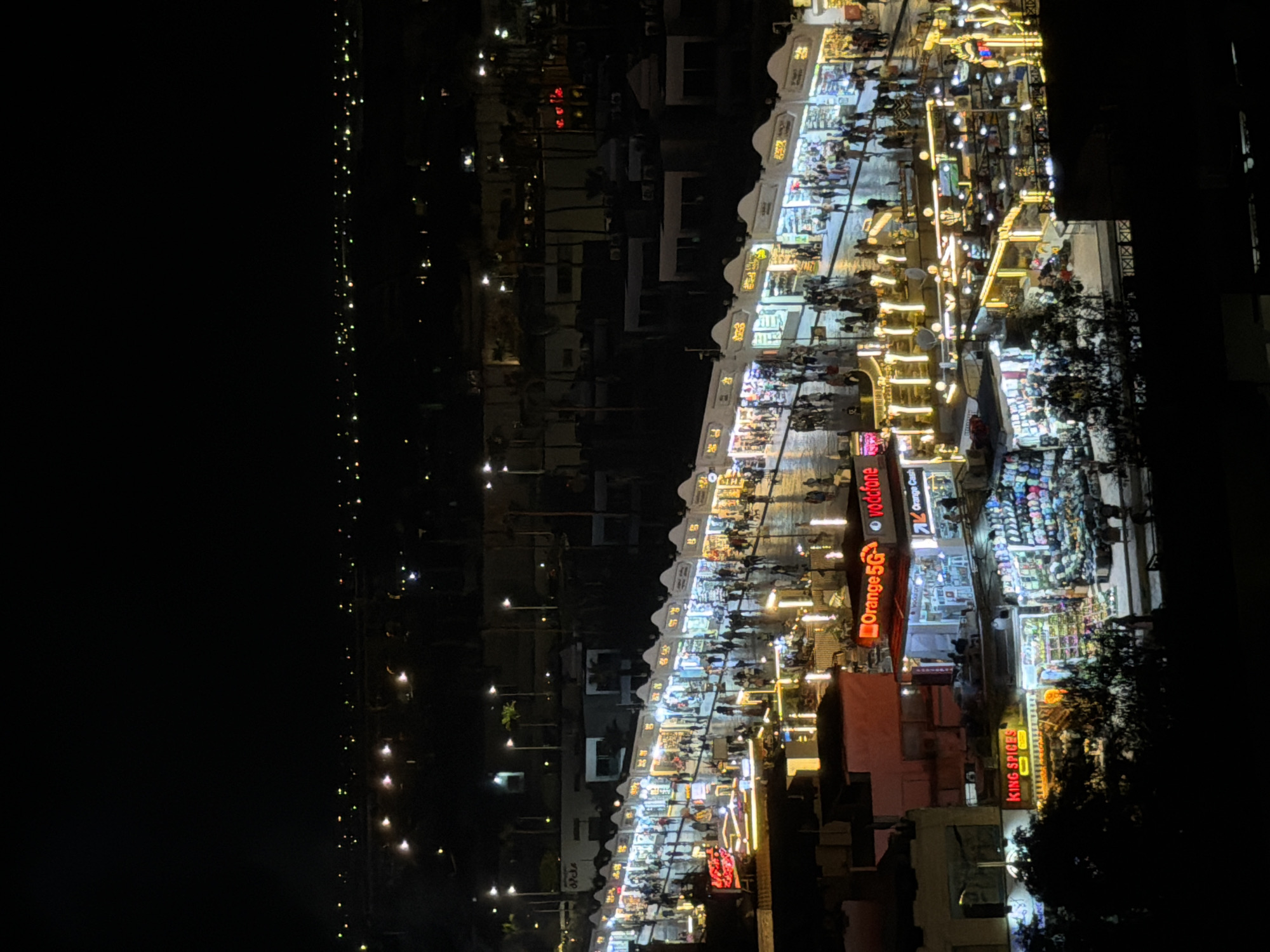 This is a nighttime aerial view of a bustling market street in Sharm El Sheikh, Egypt. The street is lined with shops and stalls, illuminated by bright, colorful lights, creating a vibrant and lively atmosphere. People are seen walking along the street, browsing the shops, and gathering in small groups. The architecture appears to be a mix of traditional and modern styles, with some buildings featuring ornate facades and others having more contemporary designs. In the background, distant city lights and palm trees can be seen, suggesting a resort town setting. Signs in Arabic and English indicate the presence of various businesses, including telecommunications providers like Orange 5G and Vodafone, as well as shops selling spices, clothing, and other merchandise. The overall scene depicts a popular commercial and social hub within the city.