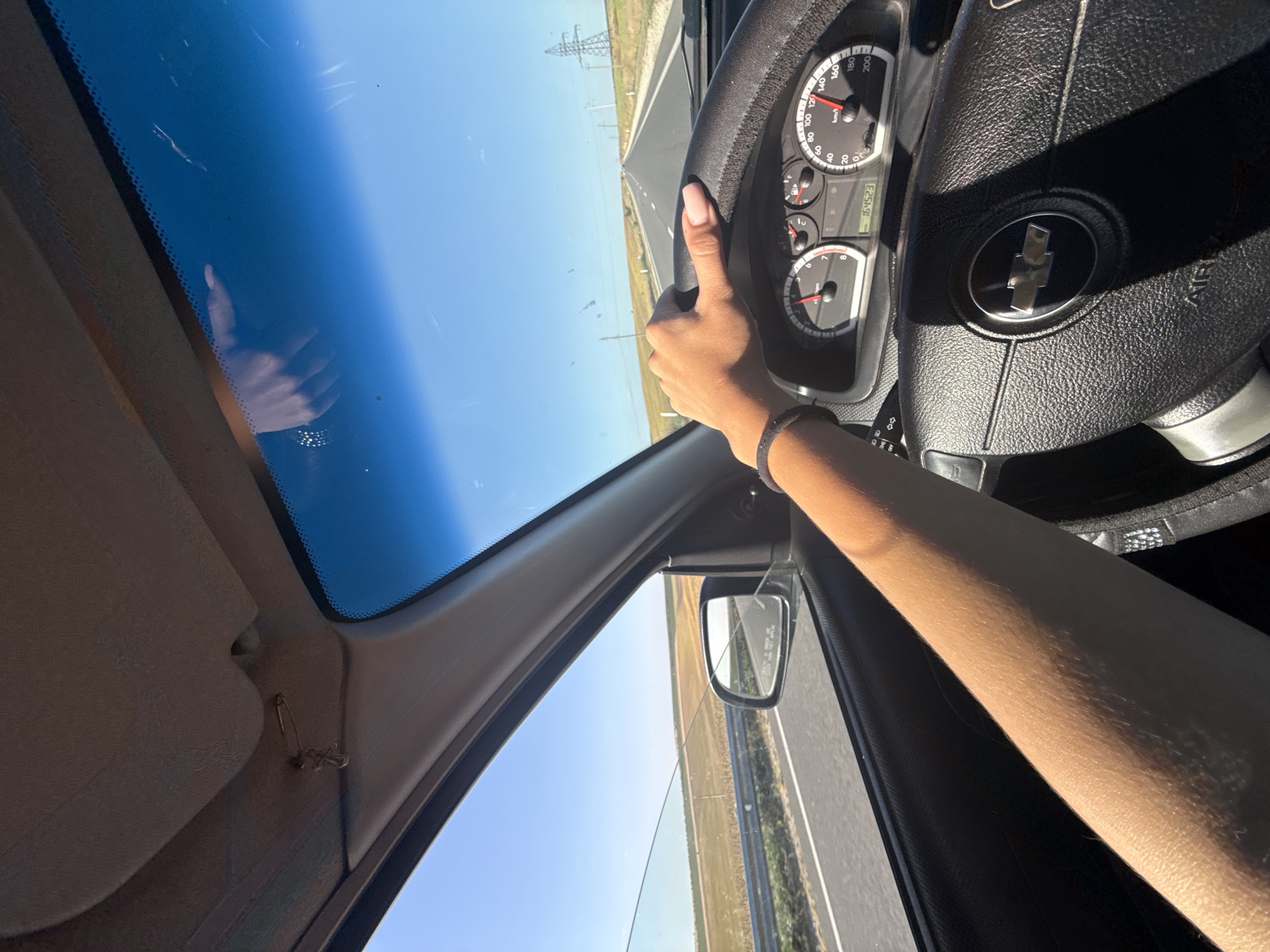 The image shows a first-person perspective of someone driving a car on a straight, paved road under a clear blue sky. The road appears to be in a rural or semi-rural area, with fields of crops and some power lines visible in the distance. The driver's right hand is on the steering wheel, and their left arm is visible resting on the car door. The dashboard of the car, including the speedometer and odometer, is also visible. A small charm hangs from the car's visor. The context of Comrat, Moldova, is not directly discernible from the visual elements of the image.