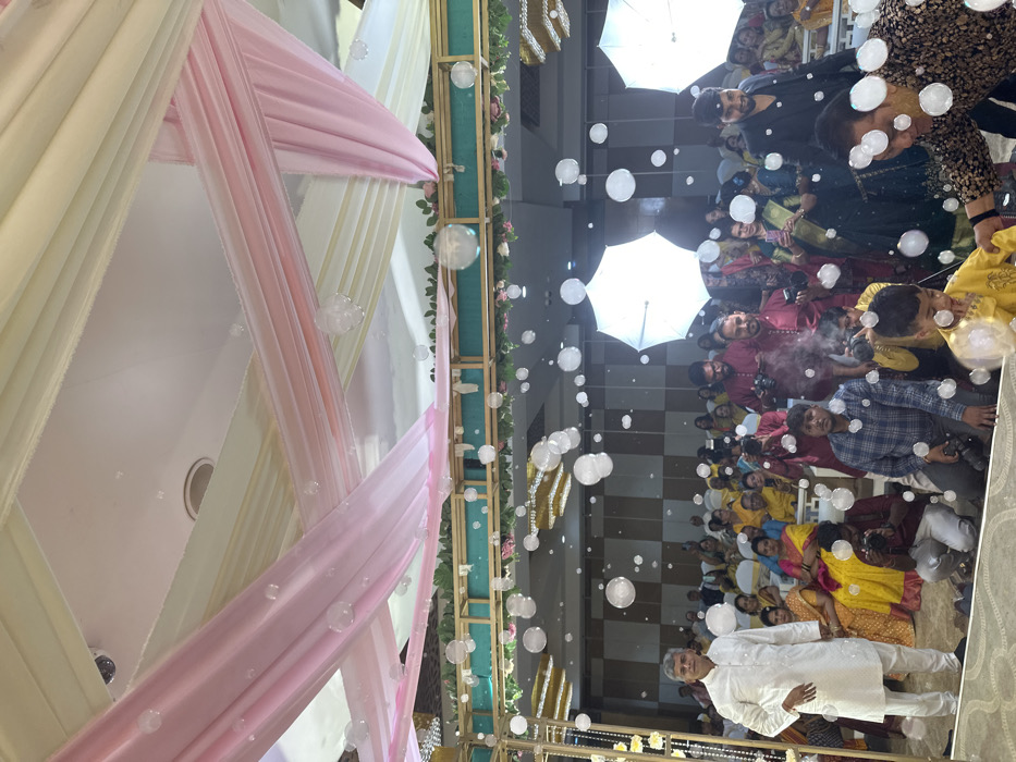 An indoor event space in Vadodara, India, is depicted from a slightly elevated perspective, showcasing a celebratory atmosphere. Numerous transparent bubbles of varying sizes are suspended in the air, falling from an unseen source on the draped ceiling. The ceiling is adorned with alternating light pink and off-white fabric drapes, secured by a golden-colored framework embellished with foliage.
In the mid-ground, a diverse group of individuals is present. To the left, a man in a white kurta pajama stands erect, looking towards the viewer. Central to the image, several men, some holding professional cameras, are kneeling or standing. One man in a maroon garment is operating a device emitting white vapor, contributing to the atmospheric effects. A young boy in a yellow kurta is visible in the lower right foreground, engaging with the bubbles. To the far right, another man in a patterned dark shirt is partially visible, leaning forward.
In the background, a large crowd of attendees, primarily women dressed in various colors of traditional Indian attire, including many in yellow or gold, are seated or standing, observing the scene. Two large, white, illuminated umbrella-like objects, likely photographic diffusers or decorative elements, are visible behind the main group of people. The overall scene suggests an event involving photography, special effects, and a gathering of guests.