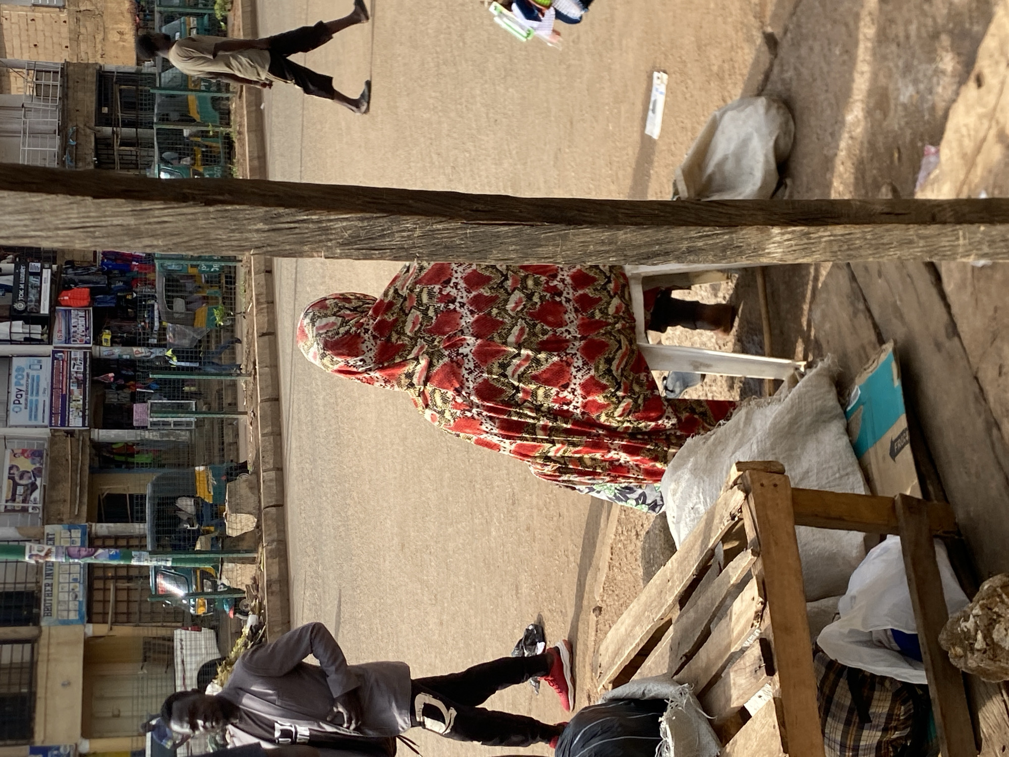 The image captures a lively street scene in Bukuru, Nigeria, during a bright, sunny day. The perspective is slightly elevated, looking down upon the activity.
In the immediate foreground, a person is seated on a white plastic chair, draped in a distinctive red, white, and black patterned fabric, facing away from the camera towards the street. Beside them, another individual stands, dressed in a grey long-sleeved top, dark trousers, and bright red sneakers. Near these figures are wooden pallets or crates, and several large sacks (one black, others light-colored), hinting at storage or market goods. A thick wooden beam or plank extends horizontally across the mid-ground, acting as a visual separator.
Further along the dusty street, another person in a light-colored shirt and dark shorts walks away from the viewer. The road appears unpaved or lightly graveled, illuminated by strong sunlight that casts distinct shadows.
The background reveals a row of buildings housing shops or market stalls. Signs are visible on some storefronts, with partial text reading "TOK M LUXURY," "Pay POIN...", and "POS" on a blue sign. Two small, enclosed three-wheeled vehicles, commonly known as auto-rickshaws or keke-napep, are parked or moving on the street. The overall atmosphere suggests a bustling commercial area, capturing a slice of daily life.