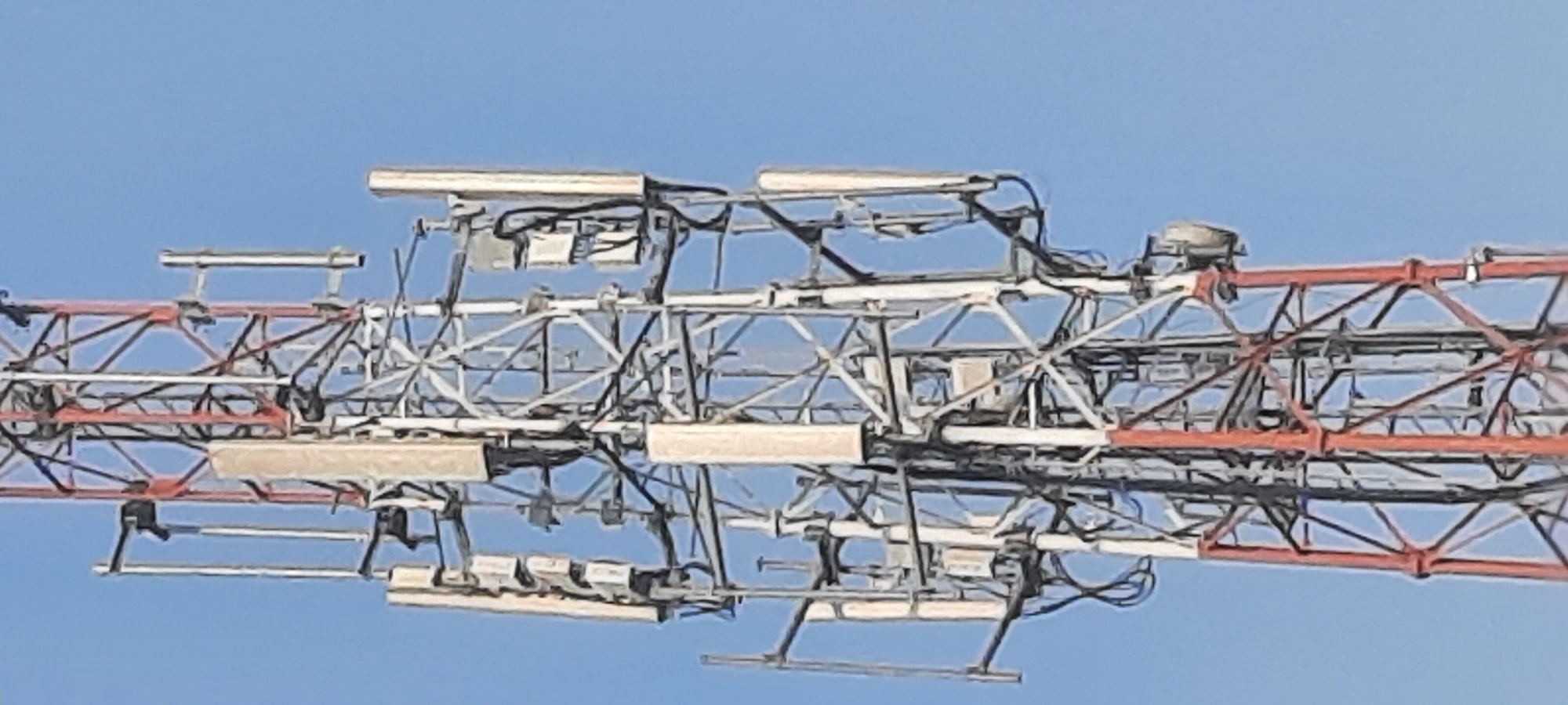 The image shows a close-up, low-angle view of a cellular tower against a clear blue sky. The tower is a lattice structure with red and white painted sections, indicating it is likely in Lodoyo, Indonesia, where such towers are common. The tower is equipped with multiple antennas, which are rectangular and light-colored, mounted on various arms. Cables are visible running along the structure. The overall impression is of industrial infrastructure against a natural, unobstructed background. There are no people or animals visible in the image, and the focus is solely on the tower and its components. The lighting suggests it is daytime, with no shadows indicating the position of the sun.