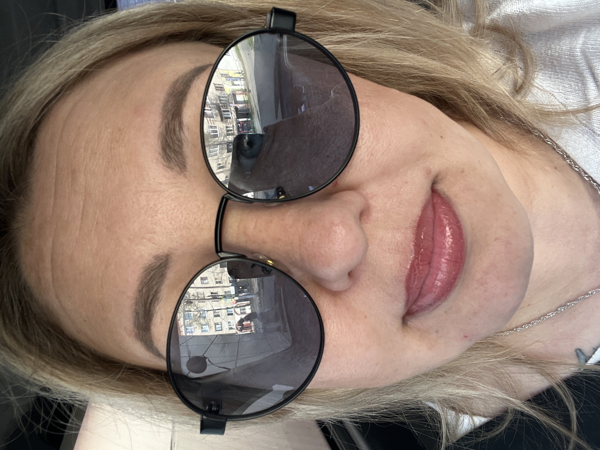 A woman with blonde hair is wearing sunglasses that reflect buildings and a street in Chaikovsky, Russia. The sunglasses obscure her eyes, but the reflection shows a city scene with multi-story buildings. She has fair skin and is wearing lip gloss. A silver chain is visible near her neck. The background is a mix of black and white clothing. The photo appears to have been taken indoors.