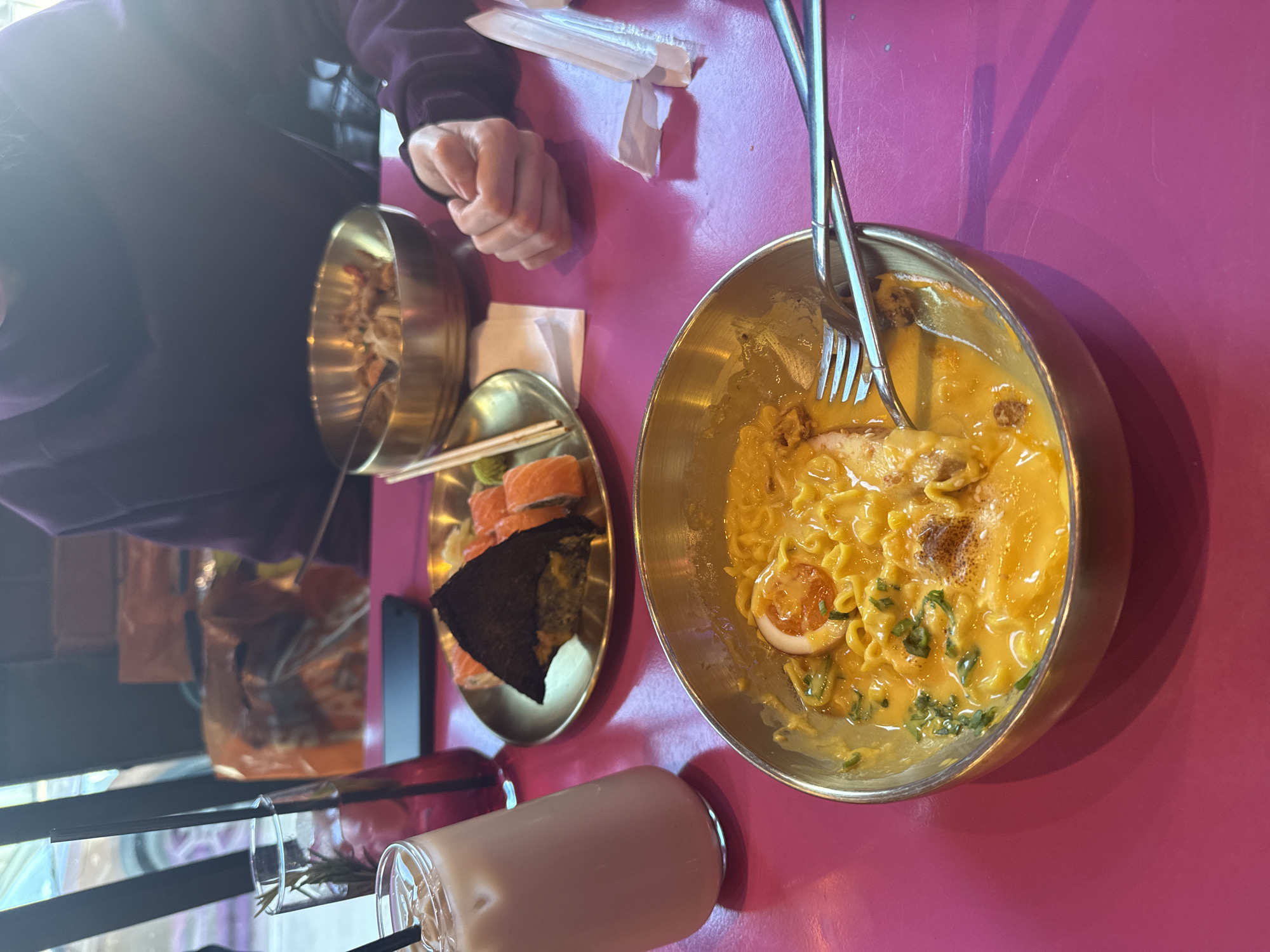 A high-angle, medium shot depicts a dining table with food and drink. The table is pink. On the right, a golden bowl contains ramen with a creamy yellow broth. Forks are positioned in the bowl. To the left of the ramen, a small golden plate holds sushi rolls and a triangular piece of nori seaweed. A golden bowl, partially visible on the left, appears to hold a dish with small pieces. A tall glass with a straw contains a light brown beverage. A person wearing a dark purple garment is visible at the top left. The hand of the person is visible near the sushi. The background is blurred. The image was taken during daylight. It is not possible to determine the location in the city of Moscow, Russia from this image.