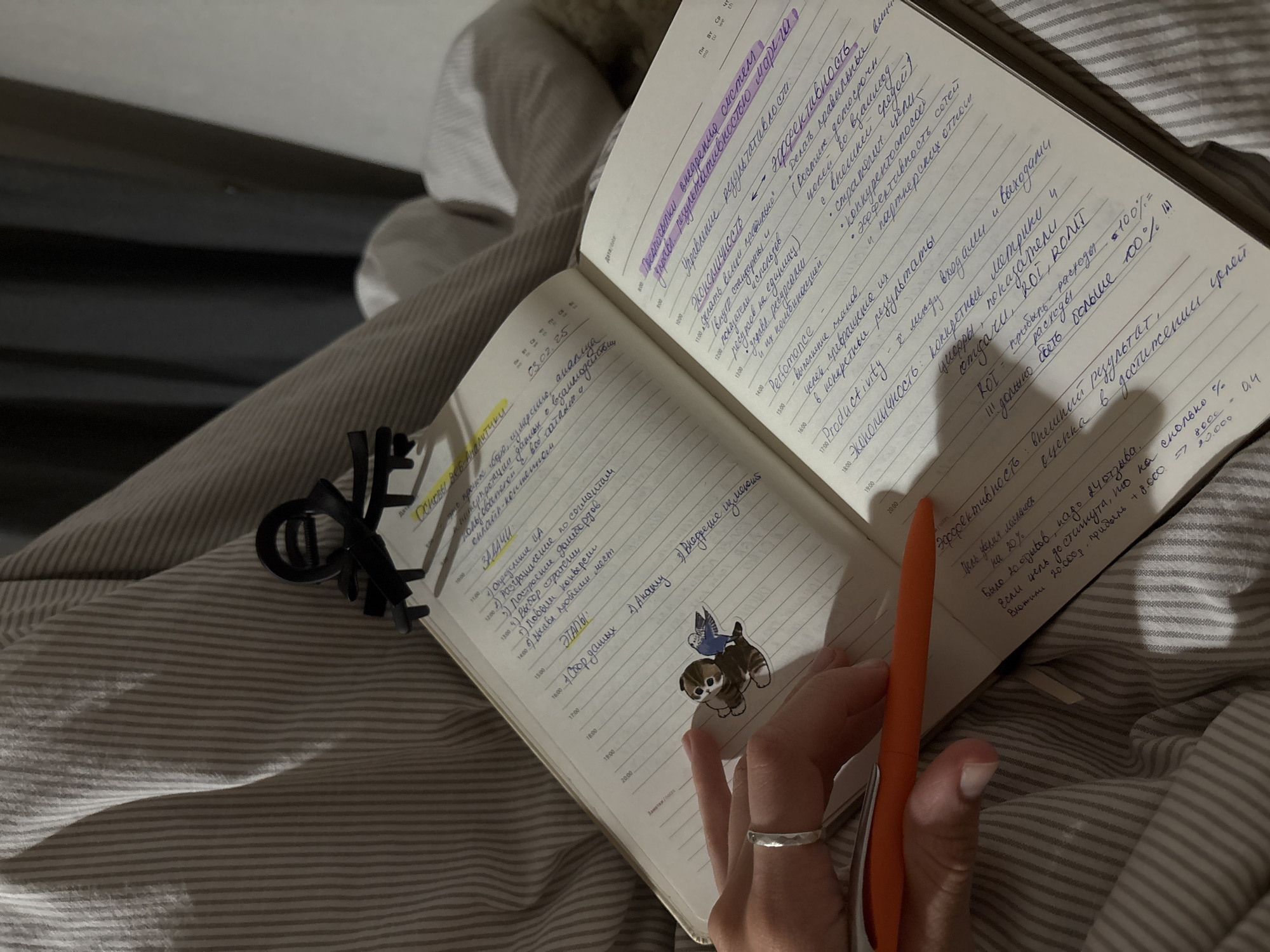 The image depicts an open notebook with handwritten notes in Cyrillic script, suggesting a personal diary or planner. The notebook is placed on a striped fabric, possibly a bedsheet or blanket, in a dimly lit indoor environment. A hand with a silver ring is visible, holding an orange pen, poised to write. A small sticker of a kitten with a bird on its back is affixed to the left page of the notebook. The overall impression is one of quiet study or contemplation. There are no discernible indicators suggesting a specific location in Perm, Russia.