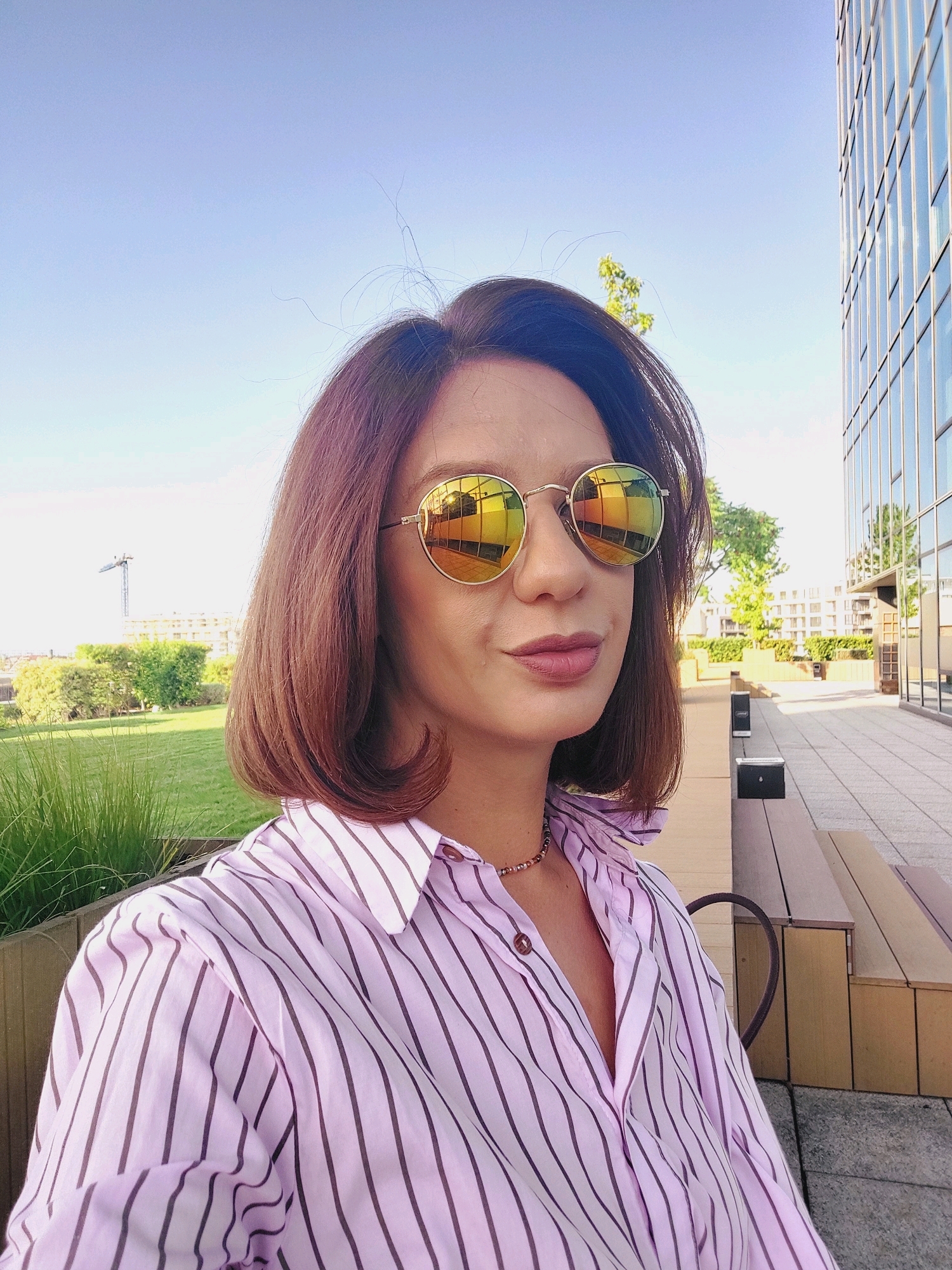 A woman with short, brown hair and sunglasses is taking a selfie outdoors. She is wearing a pink and purple striped button-up shirt and a beaded necklace. The background shows a modern building with glass windows, a green lawn, and a construction crane in the distance. The location is an outdoor terrace in Sofia, Bulgaria, with benches and paved ground. The lighting suggests it is daytime.