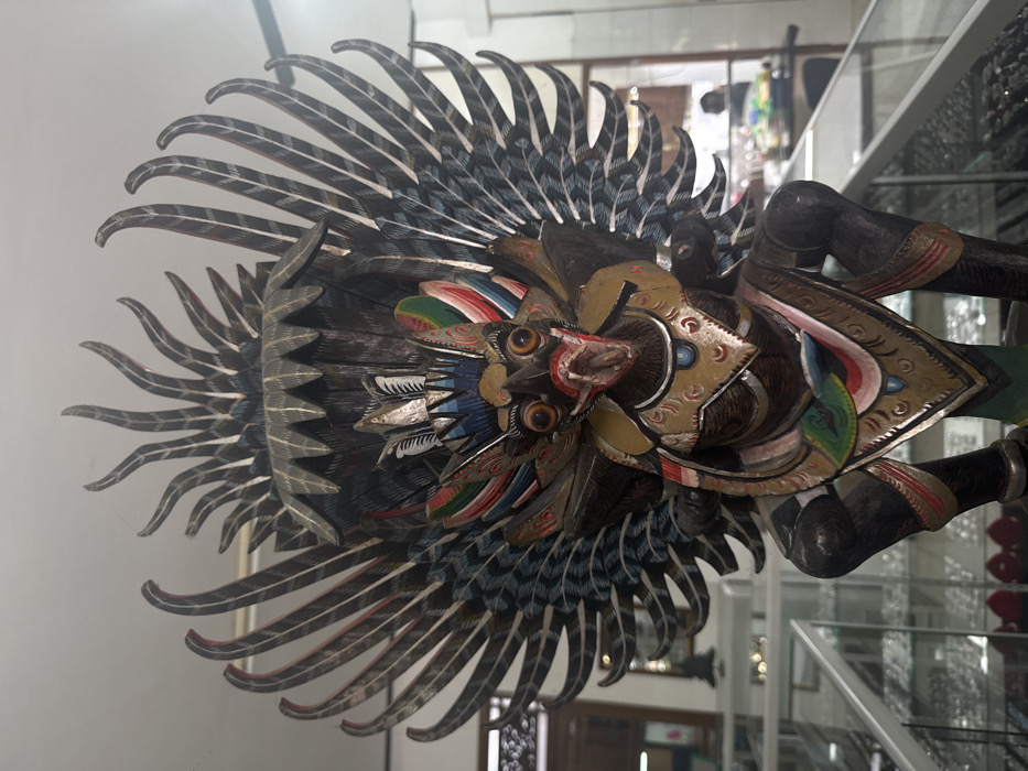 A vibrantly colored, carved wooden figure, possibly a Garuda representation, dominates the frame. It features a bird-like face with a beak, surrounded by ornate headdress and wing-like structures. The sculpture has a dark, detailed plumage with gray and black feathers accented with splashes of red, blue, and green. The figure appears to be displayed within a glass case, the surrounding environment hinting at a shop or museum setting. The piece is intricate and detailed.