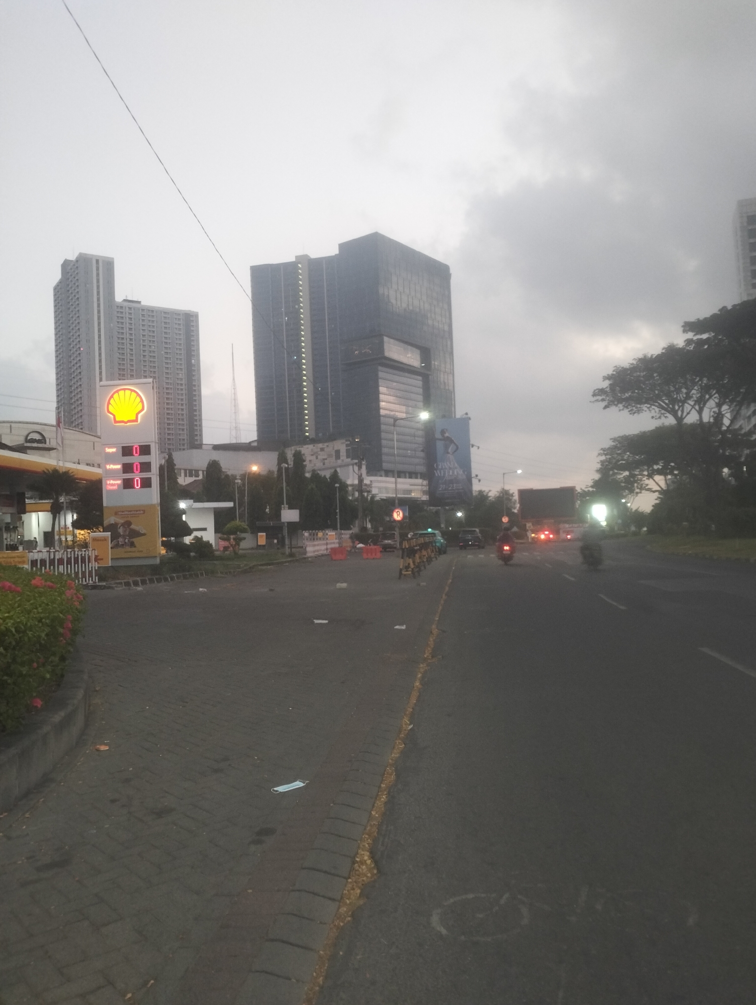 The image depicts a street scene in Surabaya, Indonesia, likely near a commercial or business district, as indicated by the presence of modern high-rise buildings and a Shell gas station. The sky is overcast and the lighting suggests it is either early morning or late afternoon, with a generally hazy atmosphere.
In the foreground, a paved road is visible, with a gray brick sidewalk to the left, bordered by a low concrete barrier and some flowering bushes. A discarded surgical mask lies on the brickwork. The Shell gas station sign, illuminated and featuring the brand's yellow shell logo, stands prominently on the left side of the image. A digital price display shows "0" for "Super" and "V-Power Diesel." Further back, a large billboard is visible on the side of a building.
The street itself appears to be a multi-lane road, with a few vehicles present. Several motorcycles are in motion, with one rider visible in the middle ground and another in the right background. A truck or larger vehicle with illuminated taillights is also on the road. The road is marked with white lines, and a yellow line runs along the edge of the sidewalk.
In the background, imposing high-rise buildings dominate the skyline. One building, a dark glass skyscraper, has a distinct angular design and lights visible on its facade. To its left, another cluster of taller, lighter-colored residential or office buildings is visible. Further in the distance, a slender tower, possibly a radio or cellular tower, can be seen. Trees line the right side of the road, adding a touch of greenery to the urban landscape. The overall impression is one of a busy, modern city under a subdued sky.