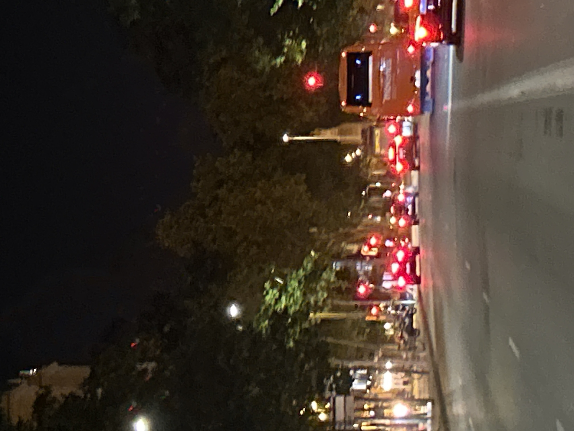 The image shows a nighttime street scene in Barcelona, Spain, featuring heavy traffic on a multi-lane road. The road stretches into the distance, lined with trees. Traffic lights and headlights create a string of red and white lights. There's a prominent orange bus among the vehicles. On the left side of the image are dark trees and a few buildings illuminated by streetlights. A street lamp is visible in the middle of the road and trees. The sky is dark, suggesting it's late at night.