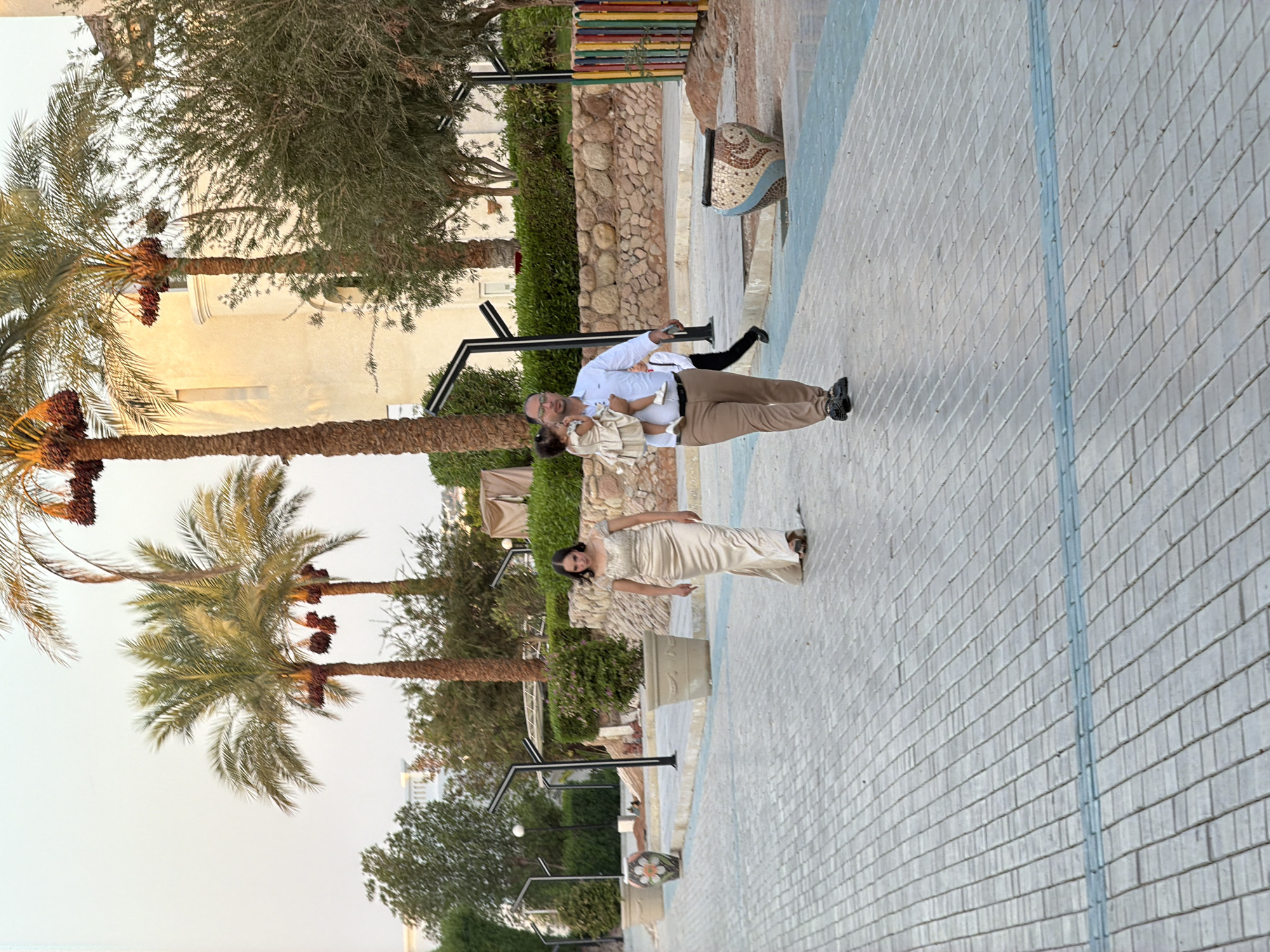 The image shows a paved walkway with a family walking. A woman in a long, gold dress is walking towards the camera on the left. On the right, a man in a white shirt and tan pants is carrying a young child. The background features palm trees, buildings, and a stone wall. The scene appears to be in a resort or public area in Sharm El Sheikh, Egypt, given the presence of palm trees and the architectural style. The sky is a pale, overcast color, suggesting either early morning or late afternoon. The walkway is made of gray pavers with a light blue inlaid strip. Decorative pots and a colorful fence are visible on the right side of the image.