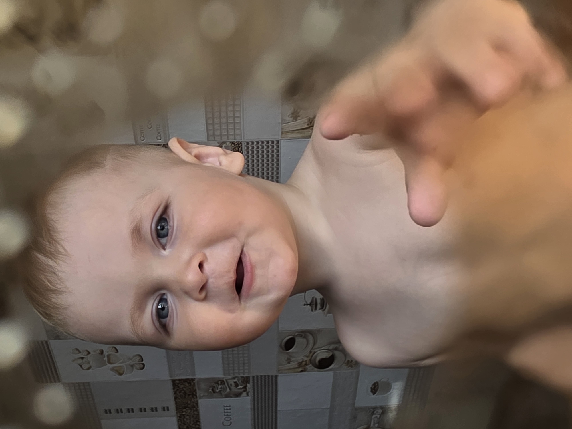 The image displays a rotated, close-up portrait of a young baby, likely a toddler, in an indoor setting in Chaikovsky, Russia. The baby has fair skin, light-colored hair, and bright blue eyes, and is depicted shirtless. Its expression is wide-eyed and open-mouthed, conveying curiosity or a playful mood, as it looks directly at the camera.
In the foreground, one of the baby's hands is heavily blurred, reaching towards the camera, suggesting movement or a very close interaction with the lens. The background features tiled walls, indicative of a kitchen or dining area. These tiles display various patterns, including geometric designs of small squares and stripes, and illustrative tiles depicting coffee cups and beans. The word "COFFEE" is clearly visible on some of these background tiles.
The lighting is soft and even, focusing attention on the baby's face, while the shallow depth of field creates a blurred effect in both the immediate foreground and the background. The photograph captures a candid, intimate moment from an overhead perspective.