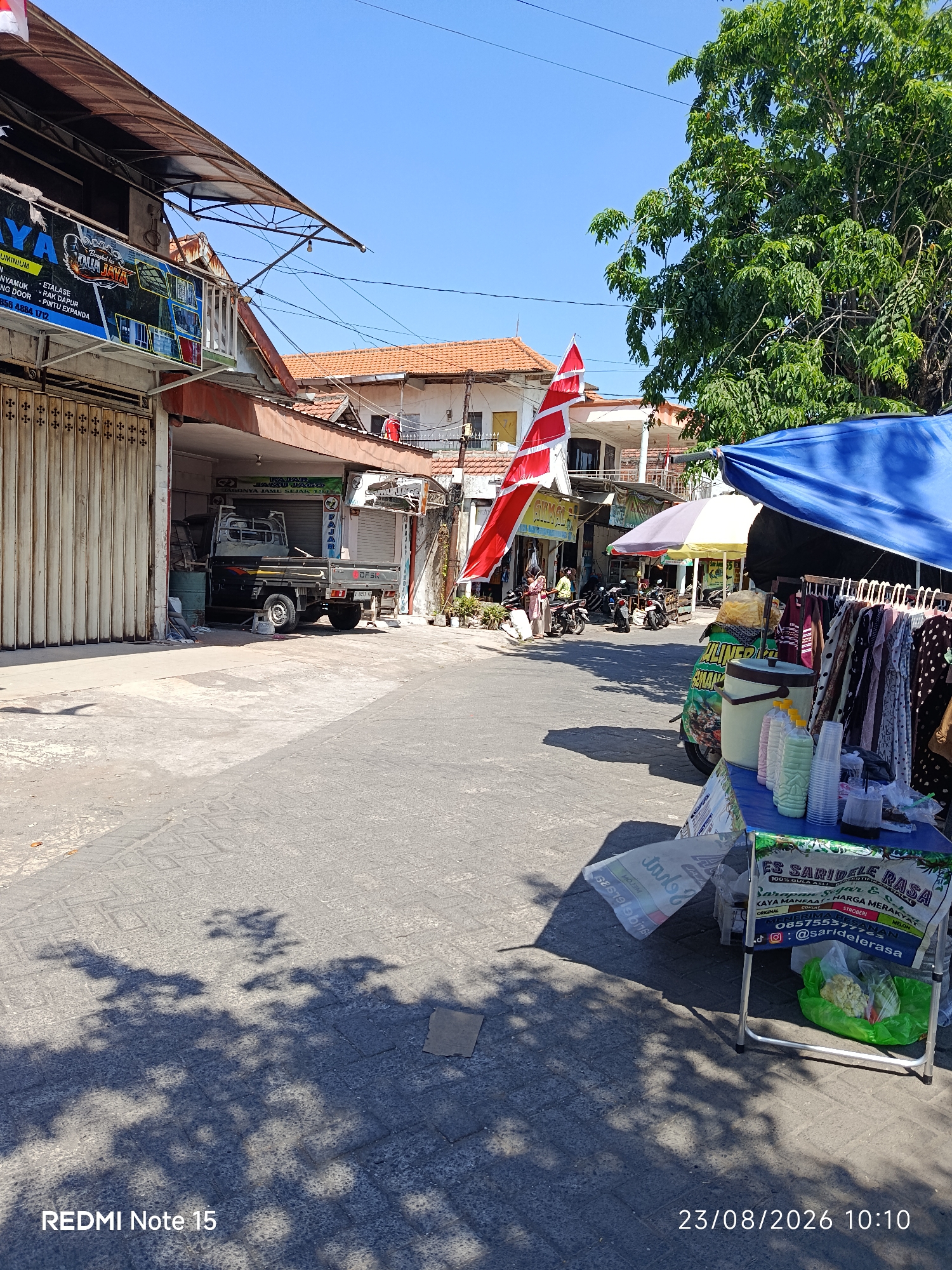 The image depicts a street scene in Keboansikep, Indonesia, on a sunny day. On the left, a business with a faded awning displays signage for "DUA JAYA ALUMINIUM." A gray pickup truck is parked partially inside a garage-like opening. The street is paved with interlocking stones. In the foreground right, a vendor stall is set up under a blue canopy, displaying clothing items for sale on a rack. Below the canopy, there are bottles of beverages, plastic cups, and a cooler. A sign on the stall advertises "ES SARIDEL rasa" with contact information. In the background, several motorcycles are parked, and people are visible near other businesses. A red and white Indonesian flag is prominently displayed on a building. Green trees provide shade on the right side of the image. The overall atmosphere suggests a bustling commercial area. The date and time stamp in the lower right corner indicates 23/08/2026 10:10.