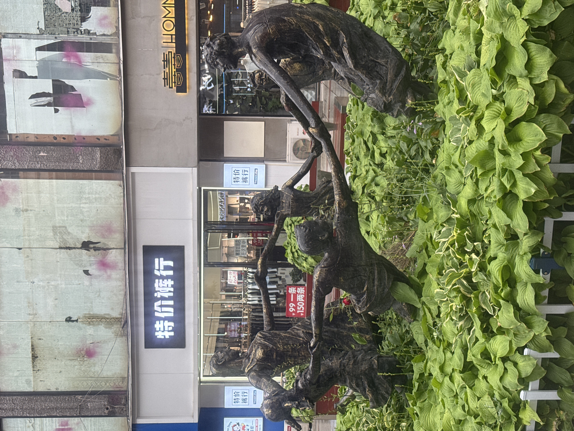 In Qinhuangdao, China, in front of a store with a sign reading "特价裤行" (Special Price Trousers) and another sign with Chinese characters and "HOMM", a bronze sculpture depicts five figures, appearing to be children and adults, holding hands in a circle amidst lush green foliage. The figures are in dynamic poses, suggesting movement and connection. The background shows a building facade with weathered panels and multiple storefronts.
