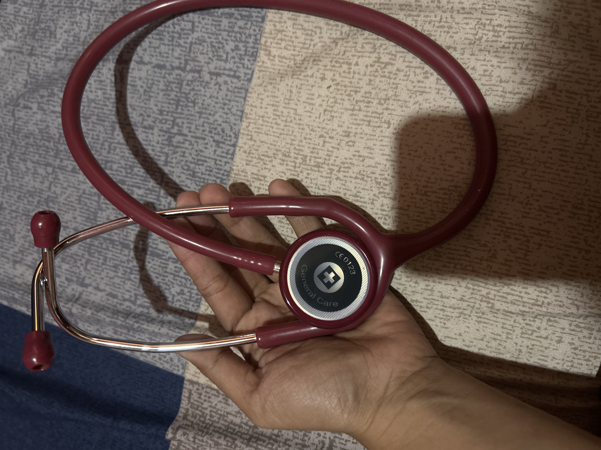 A maroon stethoscope is held by a person's hand, partially obscuring the diaphragm which has a white cross symbol and text that reads "General Care" and "CE0123". The stethoscope lies on a bed with a blue and grey patterned comforter. The scene is likely indoors in a residential setting in Depok, Indonesia.