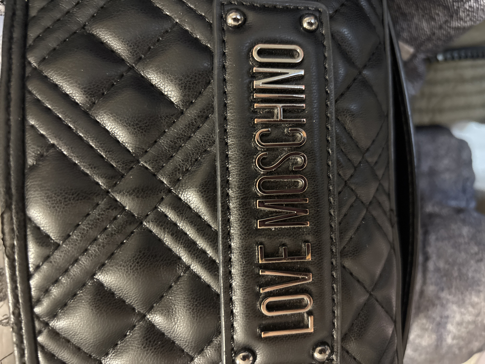A close-up, overhead shot shows a black quilted handbag with a silver metal plaque that reads "LOVE MOSCHINO." The bag is made of faux leather and has a diamond pattern stitching. The plaque is attached to the bag with two small silver rivets on either side. The background is blurred and appears to be clothing. The image does not contain any information that can be used to determine its location within Bila Tserkva, Ukraine.