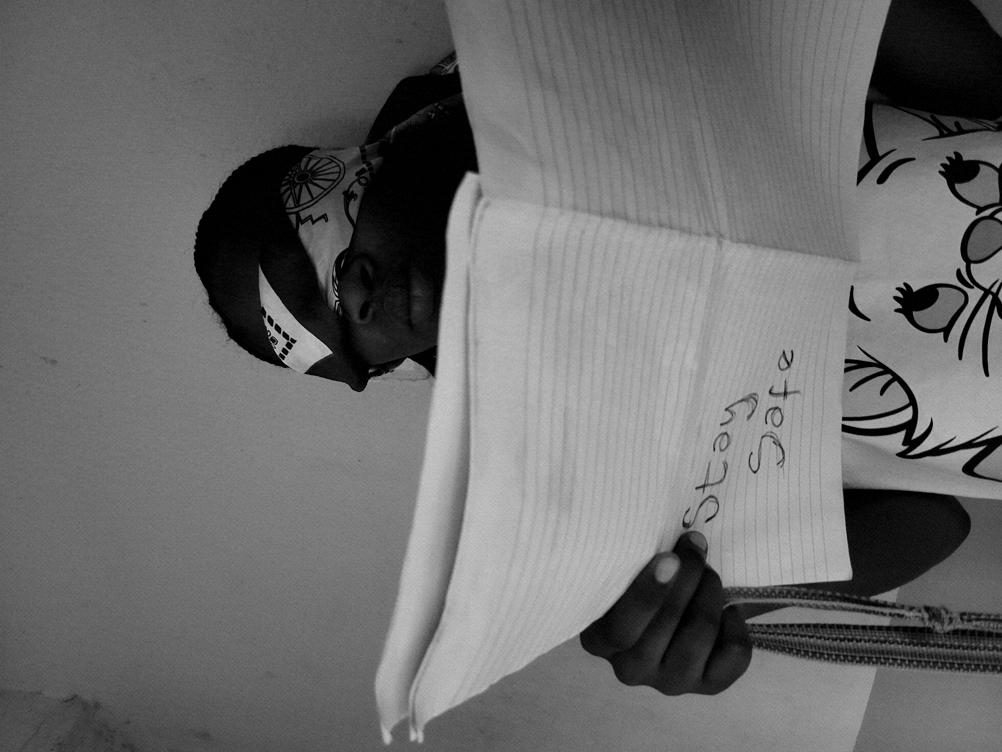 A black and white photograph shows a person holding up a piece of paper with the words "Stay Safe" written on it. The person is wearing a patterned headscarf and a shirt with a cartoon cat design. The location is Jos, Nigeria, evidenced by the person's appearance and dress. The environment appears to be indoors, with a plain wall in the background.