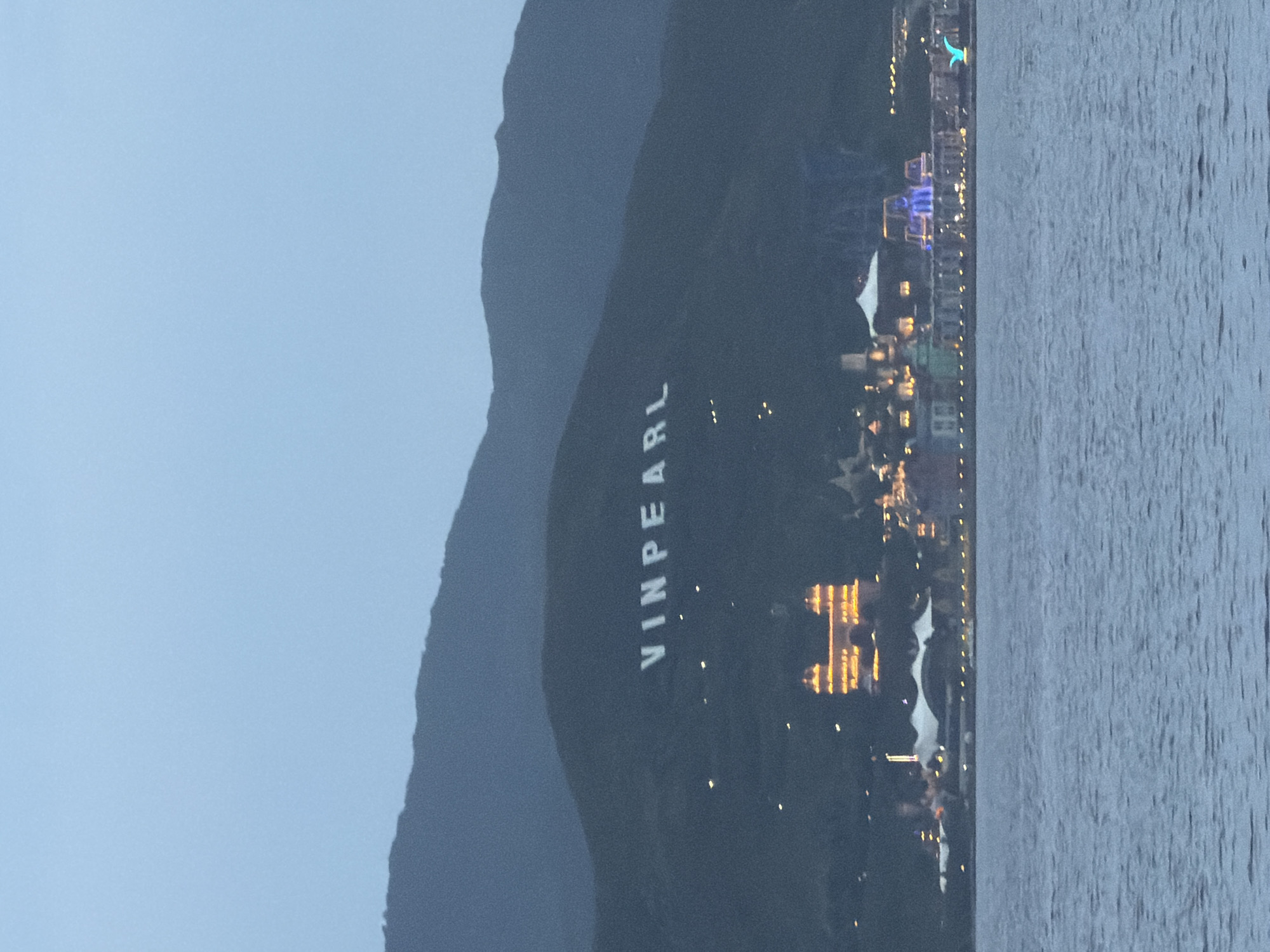 The image shows Vinpearl Land, a popular amusement park and resort located on Hon Tre Island, off the coast of Nha Trang, Vietnam. The resort is illuminated at dusk, with lights twinkling from the buildings and on the mountainside where the Vinpearl logo is emblazoned. The Vinpearl logo, in large white letters, is prominently displayed on the dark green slope of the mountain, a landmark visible from the mainland. Below the logo, a cluster of brightly lit buildings forms the resort complex, featuring a grand, castle-like structure with illuminated windows and other entertainment venues. The water in the foreground is a deep blue, reflecting the twilight sky, and the overall scene conveys a festive and enchanting atmosphere.