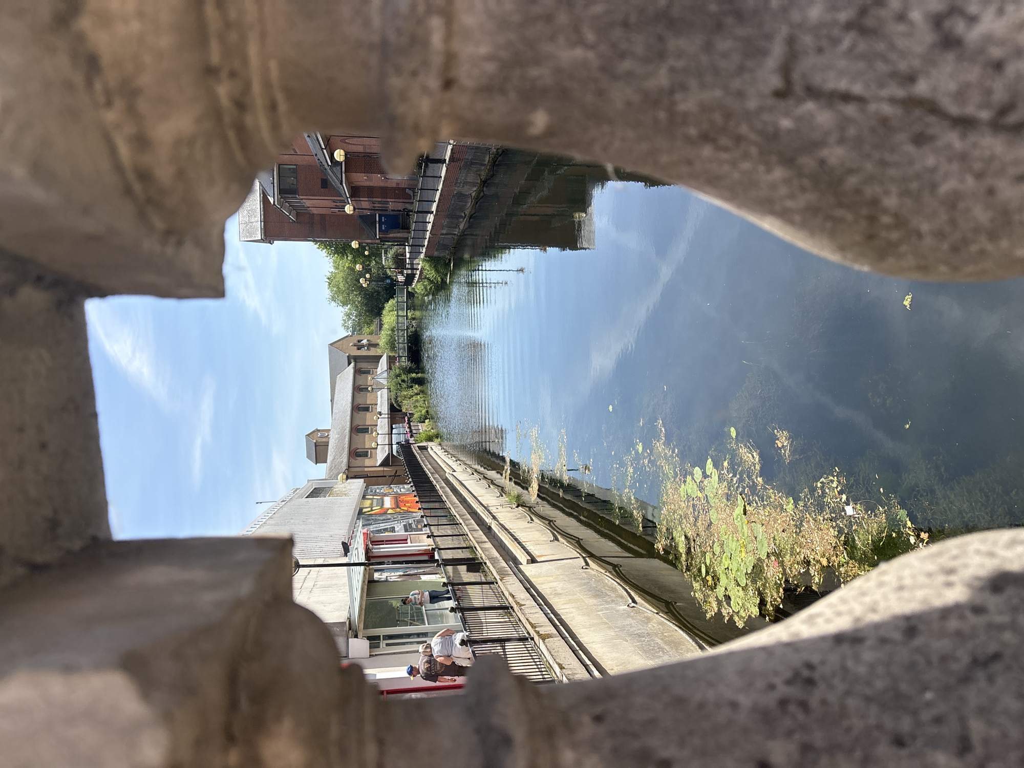 This media file depicts a canal scene, framed by rough, grey stone elements in the foreground. The perspective is from above, looking down and across the water.
The canal itself is dark and reflects the sky, with ripples on its surface. Vegetation, appearing as a cluster of green and brown aquatic plants, floats in the water on the right side of the frame. Along the left bank of the canal, a series of buildings and structures are visible. These include brick buildings with windows and rooflines, interspersed with trees. Further along the bank, a modern, multi-story building with large windows is present, with a black metal railing running along its facade.
Several people are visible on the left bank. On a walkway in front of the modern building, two individuals are standing near the railing, looking towards the canal. Another person is visible further down the walkway, seemingly walking. In the foreground, on a raised platform, two people are seated. One is wearing a dark t-shirt with "GUINNESS" visible on it.
The scene appears to be set during the daytime, with clear blue skies and sunlight casting reflections on the water. The presence of people suggests a leisurely or social setting, possibly a waterfront area with shops or cafes. The stone framing in the foreground creates a sense of looking through an opening, adding depth to the composition. The overall mood is serene and observational.