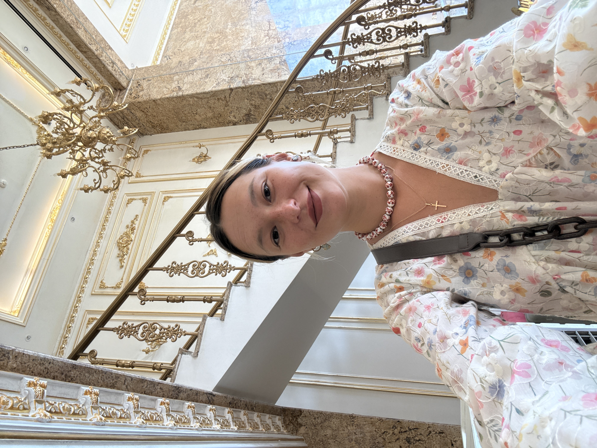 A young woman with light brown hair pulled back and freckles across her nose is taking a selfie in front of an ornate staircase. She is wearing a white floral dress with a V-neckline trimmed with lace, a beaded necklace, and a cross pendant. A dark shoulder bag with a chunky chain strap is slung across her chest. Behind her, a grand staircase with a golden railing and marble accents curves upwards. A gilded chandelier hangs from the ceiling. The background details suggest a luxurious interior, likely a hotel or a historic building. This scene is located in Da Nang, Vietnam.